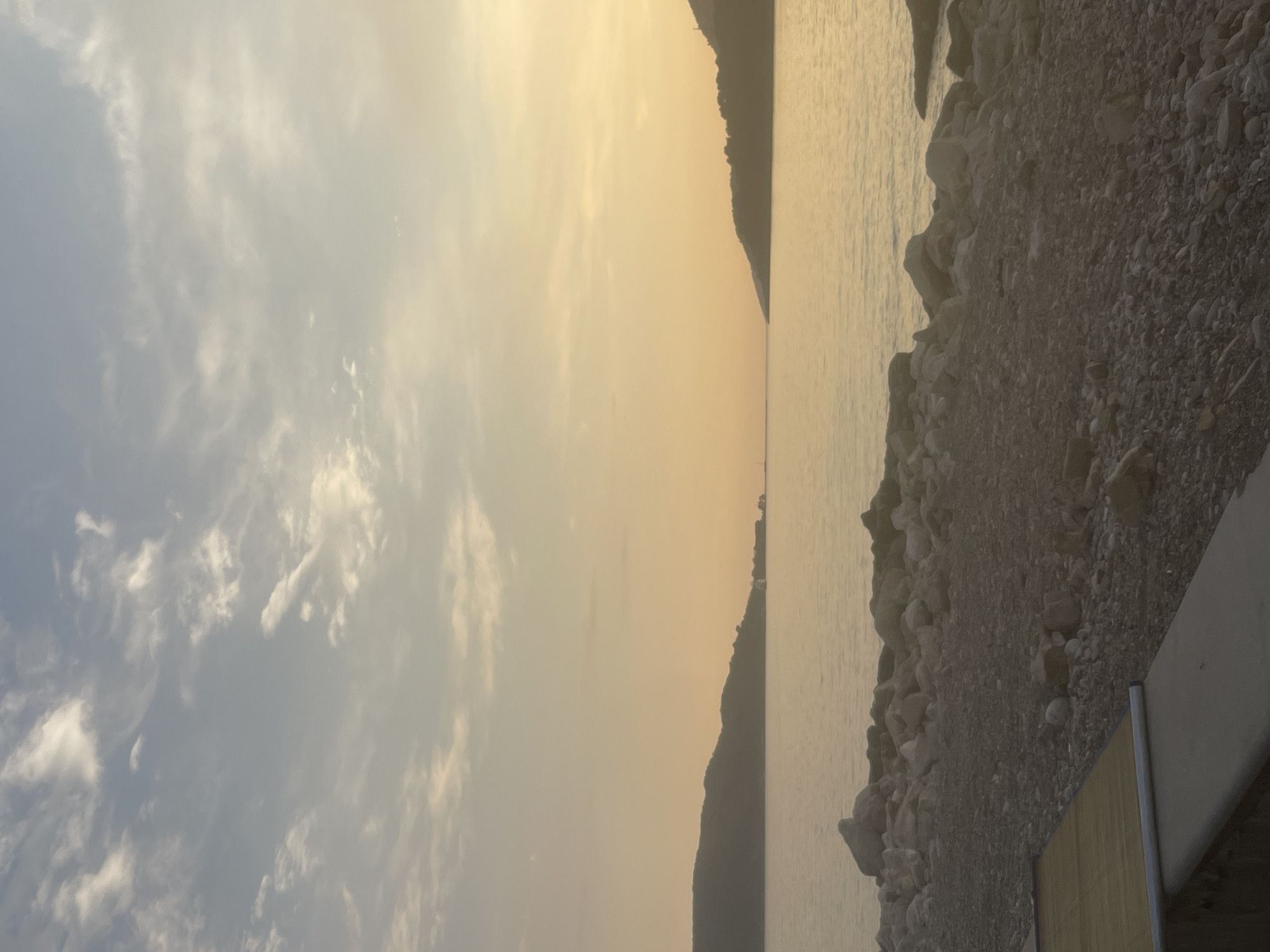 The image depicts a serene coastal scene, likely in Rafailovići, Montenegro, during what appears to be either sunrise or sunset. The sky is filled with soft, wispy clouds, illuminated by a warm, golden light that washes over the entire frame. The horizon is dominated by the silhouette of a dark, mountainous coastline, dividing the sky from the calm, reflective water.
In the foreground, to the right of the image, is a rocky shore, composed of numerous pebbles and larger stones. This natural embankment transitions into a man-made structure, possibly a part of a boardwalk or a viewing platform, with a light-colored railing and a wooden surface visible. The water itself is a muted gold and grey, smooth and still, suggesting a peaceful atmosphere.
There are no people or distinct objects of interest, apart from the natural elements and the man-made structure. The activity appears to be the passive observation of the landscape. The overall mood is tranquil and contemplative, amplified by the soft lighting and the quiet expanse of sea and sky. The time of day is clearly indicated by the quality of the light and the long shadows, if any were cast. The location is hinted at by the mountainous coastline and the presence of a coastal settlement, consistent with the geography of Montenegro. No text is visible in the image.