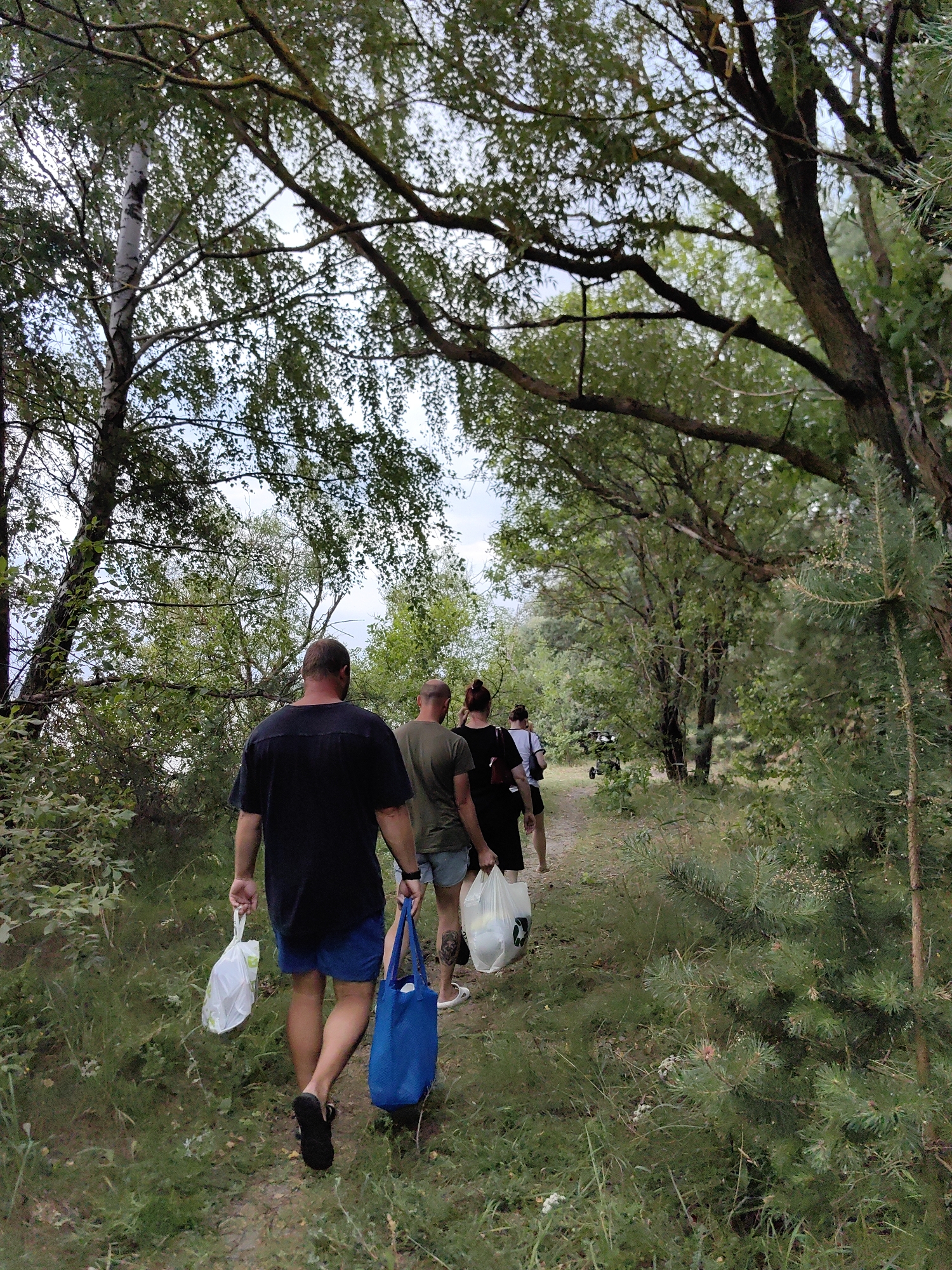 A group of four people are walking along a dirt path in a wooded area. The person in the foreground, a male, is wearing a black t-shirt and blue shorts, carrying a white plastic bag in his left hand and a blue tote bag in his right. He is walking away from the camera. Behind him, a male wearing a green t-shirt and shorts, and two females, one in a black dress and the other in a white top and shorts, are also walking along the path. They appear to be carrying white plastic bags. The path is lined with trees and vegetation, with sunlight filtering through the leaves. The scene suggests an outdoor recreational activity.