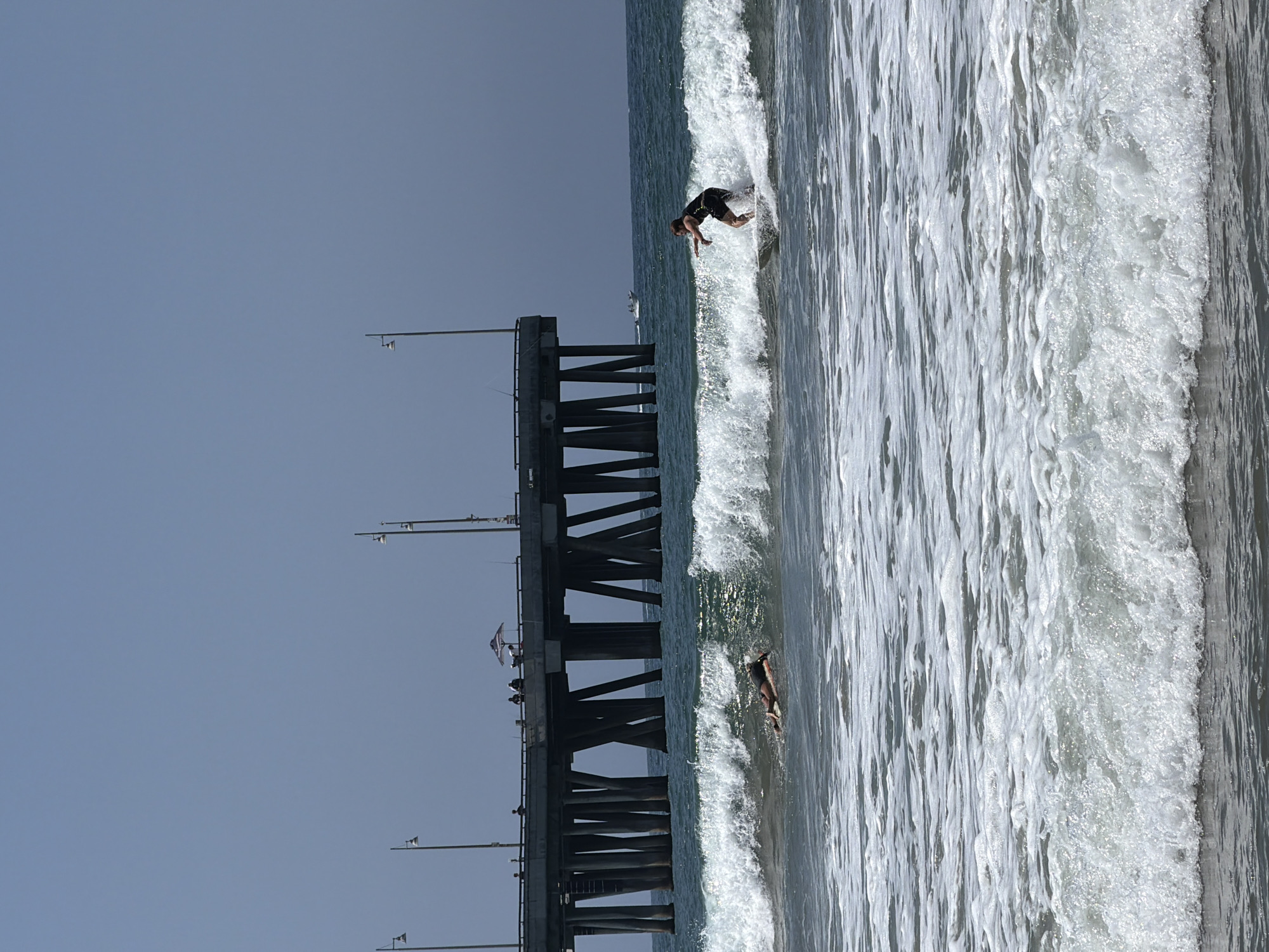The image depicts a coastal scene with a pier in the background and two surfers in the foreground. A person is body surfing in the mid-ground, lying prone on a surfboard in the breaking waves. To the right, another surfer is riding a wave, standing on their board with their arms extended for balance. The ocean is characterized by white-capped waves and foam. The pier, supported by numerous pilings, extends over the water, with a few individuals visible on its surface. The sky is clear and blue.