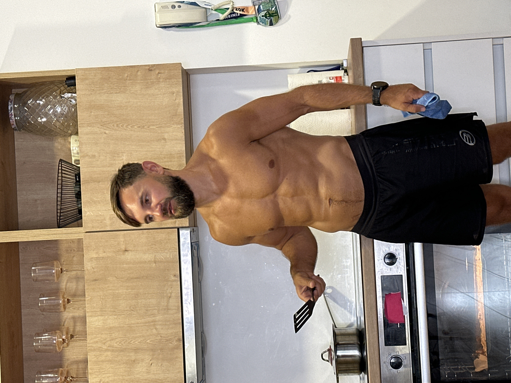 A man with short brown hair and a beard, wearing black athletic shorts, stands in a kitchen. He is shirtless, displaying defined abdominal muscles and an athletic build. In his right hand, he holds a black spatula, and in his left hand, a blue cloth. He is looking to his left. Behind him, a stainless steel stovetop is visible, with a pot on the left burner. The kitchen features wooden cabinets above the stovetop, containing glassware. A portion of an oven is also visible below the stovetop, with food inside. On the wall to the right, a white device with medals hanging from it is mounted. The scene is set indoors, within a domestic kitchen environment, possibly in Donji Pobori, Montenegro, given the context of the request.