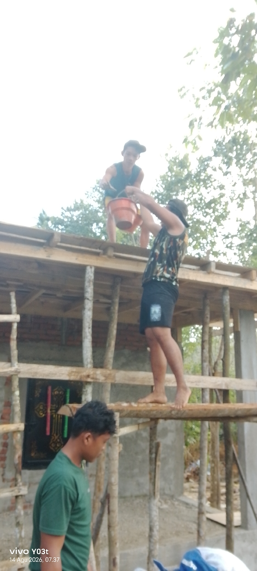 Two young men are working on an unfinished structure. The man on the left, wearing a green t-shirt, stands below and looks towards the right. The man on the right, wearing a dark tank top with a camouflage pattern and shorts, is standing on a wooden beam above the other man. He is holding an orange bucket and appears to be handing something small into it, possibly a tool or ingredient, to the man below. The structure is made of wood and bamboo, and appears to be a roof under construction. In the background, trees and foliage are visible against a bright, overcast sky. The scene is likely outdoors in a rural or semi-rural area, possibly a construction site for a home. The timestamp on the image indicates it was taken in Woiha, Indonesia on August 14, 2026, at 07:37.