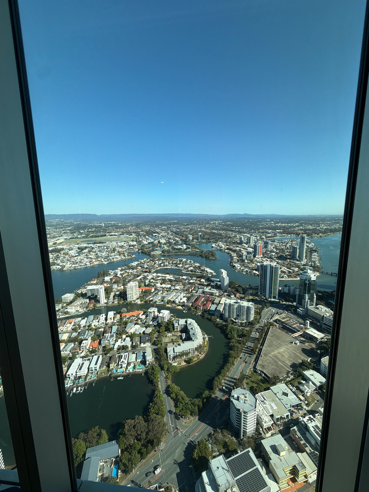 This is a high-angle, panoramic, exterior, daytime, full shot from a building overlooking a coastal city with many canals and a clear blue sky. The camera is looking out through a window, with the window frame visible on the left and right sides of the image. The city below is densely populated with a mix of high-rise buildings, apartment complexes, houses, and canals. There are many boats on the canals and scattered throughout the waterways. In the distance, there are mountains visible. The city is well-lit by the sun, indicating a clear day. The scene appears to be the Gold Coast, Australia, specifically Surfers Paradise, known for its high-rise buildings and extensive canal system. The architecture suggests a modern, resort-like environment. There are no people or discernible actions visible in the image.