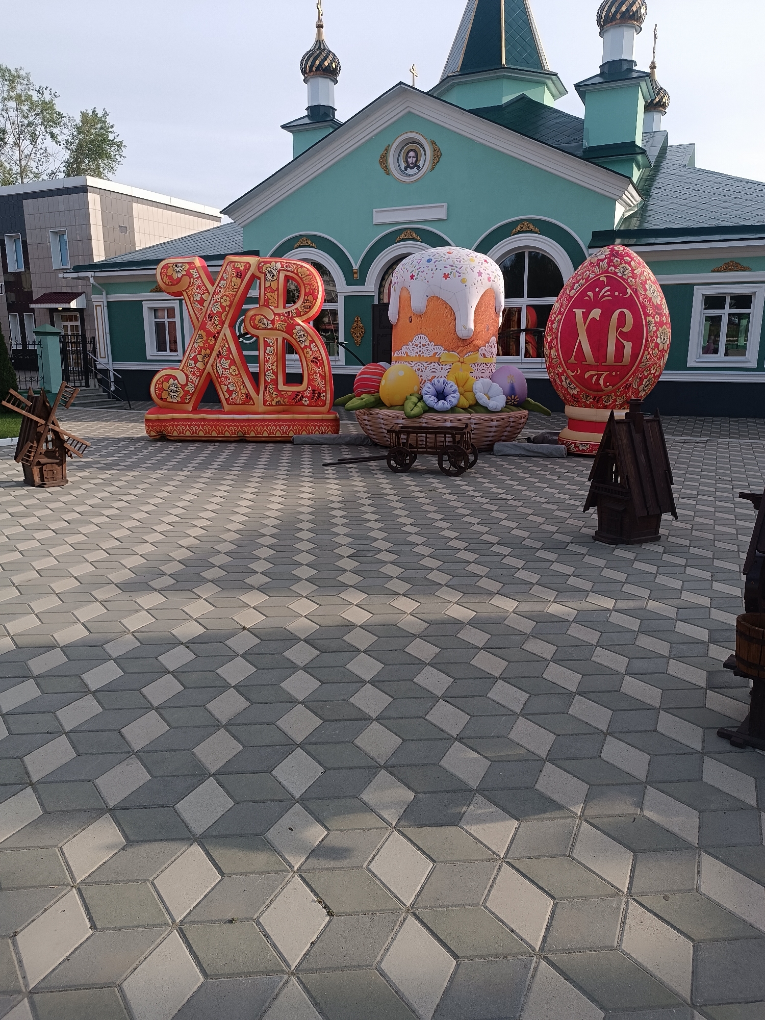 The image shows a wide-angle shot of a paved courtyard in front of a teal and green building with white trim, likely a church, in an urban location. The courtyard features grey and white interlocking paving stones in a geometric pattern. In the foreground, there is a collection of oversized Easter decorations. These include large, decorative letters "XB" in red and gold, a cake decorated with white icing and multicolored sprinkles, and a large red Easter egg with the letters "XB" on it. These items are arranged in a row behind a small wooden cart filled with artificial flowers. Additional small wooden decorative objects are scattered around the foreground. The architectural style of the building indicates a possible Russian Orthodox church with its gold domes and a portrait above the door. The sky is visible above the building, suggesting the time of day is likely morning or late afternoon. The specific city or location is not identifiable from the provided image.