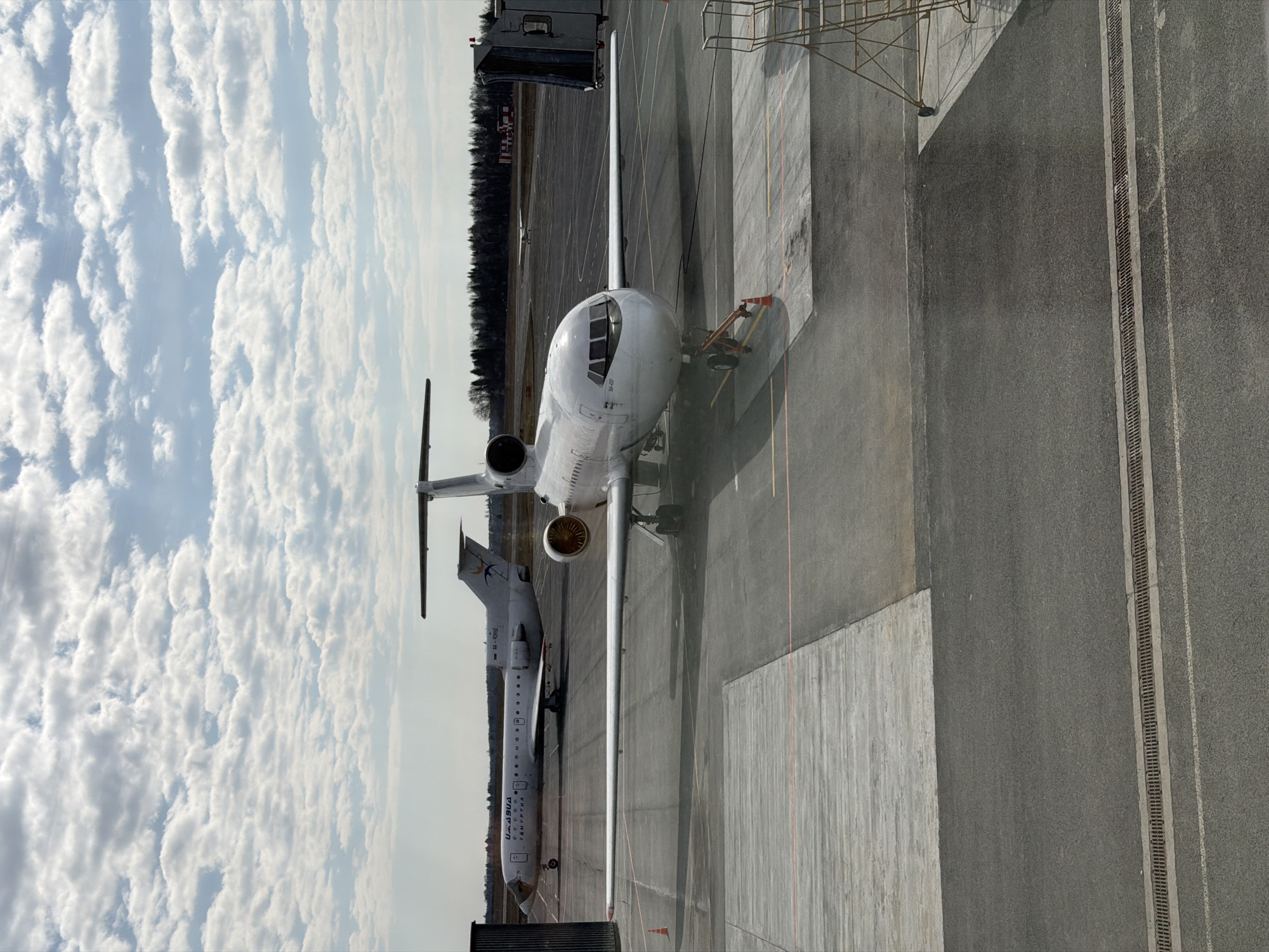 The image captures an airport tarmac scene, likely at Izhevsk, Russia, under a cloudy, daytime sky. Two white twin-engine regional jet aircraft are visible.
In the foreground, a white jet is positioned, viewed from its front-right quarter. A yellow pushback tug is connected to its nose landing gear, indicating it is either being prepared for pushback or has just been positioned. The aircraft displays the registration "RA-67123" on its nose. Its cockpit windows, engines mounted on the rear fuselage, and a portion of its wing and tail are clearly visible. A dark utility truck is partially seen behind the aircraft's left wing, and a metal ladder or scaffolding structure is situated near its right wing.
Further back and to the left, a second, similarly sized white jet is parked. Its tail is adorned with a bird logo, and the Cyrillic text "ИЖАВИА" (IZHAVIA) is distinctly visible along its fuselage, confirming the context of a Russian regional airline. Part of its registration, "RA-67...", is also discernible.
The tarmac consists of various shades of grey asphalt and concrete sections, marked with orange and white lines. A drainage grate runs along a section of the pavement. Some areas of the ground appear wet, possibly from recent rain or de-icing. A dense line of trees forms the horizon in the background, beneath a sky filled with scattered white clouds. No people are clearly visible in the frame.