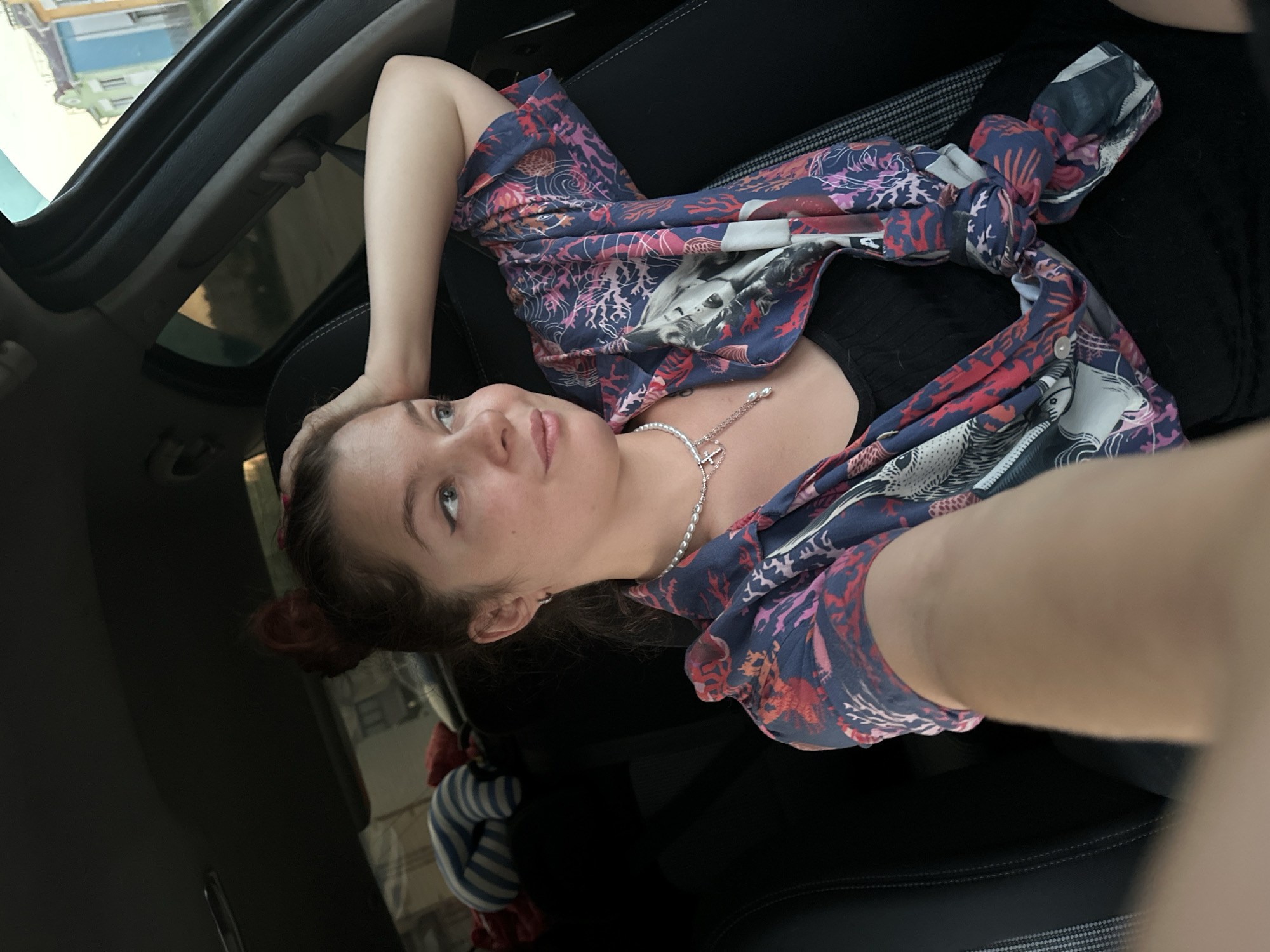 A young woman with auburn hair pulled into a bun, is sitting in the driver's seat of a car, resting her head on her hand and looking to the right. She is wearing a black tank top and an open, patterned button-up shirt with blue, red, and pink designs, possibly depicting marine life. A pearl necklace with a silver charm is visible around her neck. The interior of the car, including the black leather seats and part of the dashboard, is visible. Outside the car window, a street scene is visible with buildings of various colors, including a blue and a green building, suggesting an urban environment. The lighting suggests it is late afternoon or early evening. The presence of the buildings with their architecture and coloration could indicate the city of Vladimir, Russia.