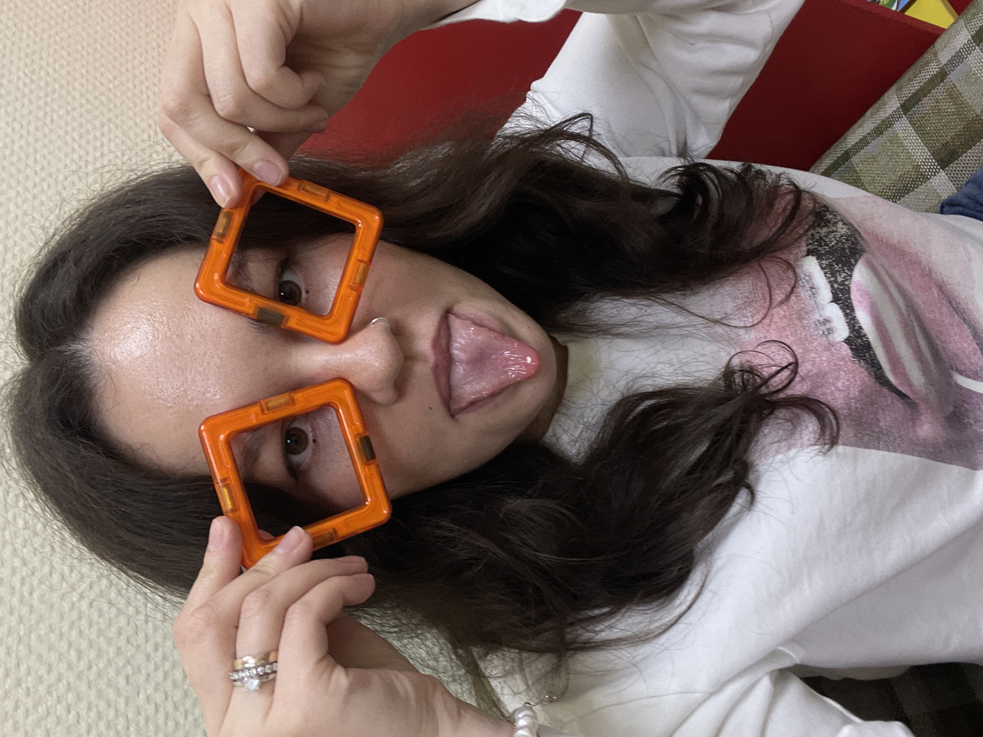 A young woman is captured in a playful and casual pose, lying down indoors. She is holding two vibrant orange square magnetic tiles over her eyes, using them as oversized, blocky frames, giving the impression of humorous glasses. Her dark hair is spread around her head, and she has her tongue playfully sticking out. She wears a white long-sleeved shirt, which features a graphic design on the front, although no specific text is legible. On her left ring finger, multiple rings are visible, including one with a prominent clear stone, and a pearl bracelet adorns her wrist. The background indicates an indoor setting, with a red cushion or pillow and a plaid fabric item, likely a blanket or another cushion, behind her. The surface she is lying on is a light-colored, textured material, possibly a carpet or rug. The image conveys a lighthearted and informal mood. This scene is set in Izhevsk, Russia.