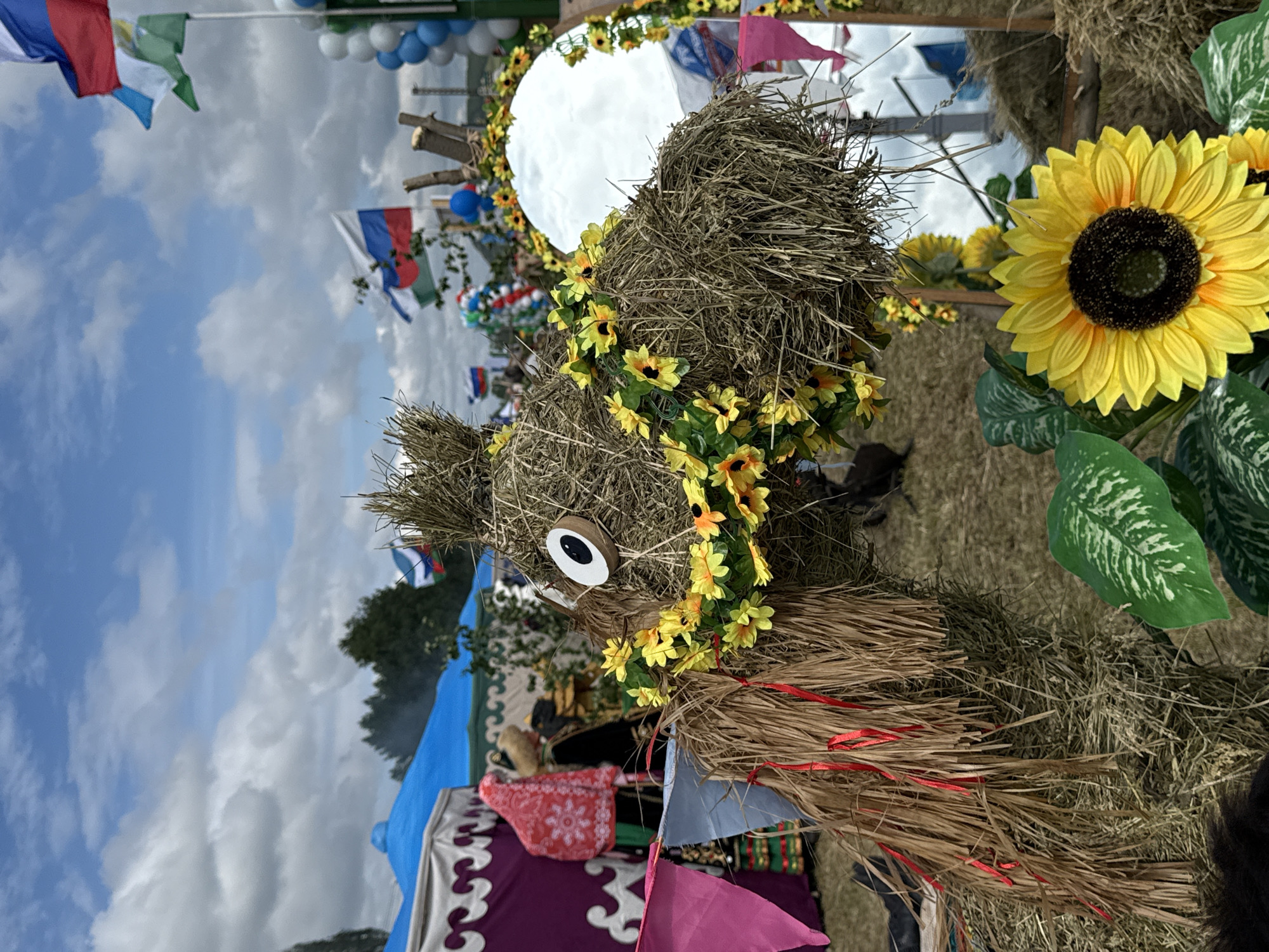 This is a medium-shot, eye-level, outdoor photograph taken on a cloudy day. The foreground is dominated by a straw sculpture of an animal, possibly a camel or horse, adorned with a garland of yellow artificial sunflowers and red ribbons. The sculpture has a large, cartoonish eye made of wood and black felt. To the right of the sculpture is a round mirror reflecting the sky and surrounding scene, framed by straw. In front of the mirror, a large, artificial sunflower is placed.
In the middle ground, there are several tents with colorful fabrics, including a purple and white patterned yurt-style tent and a blue tent. People are visible in traditional clothing, one wearing a red headscarf and a decorated outfit, and another in darker clothing with a fur-trimmed hat. Russian flags are visible on flagpoles in the background. Balloons in white, blue, and green are also present, suggesting a festive occasion. The ground is covered in dry grass and straw.
The background shows more tents, trees, and the sky with scattered clouds. The overall impression is of an outdoor festival or fair held in a rural or semi-rural setting, potentially in or near the village of Старое Субханкулово, Russia, given the Russian flags and cultural elements.