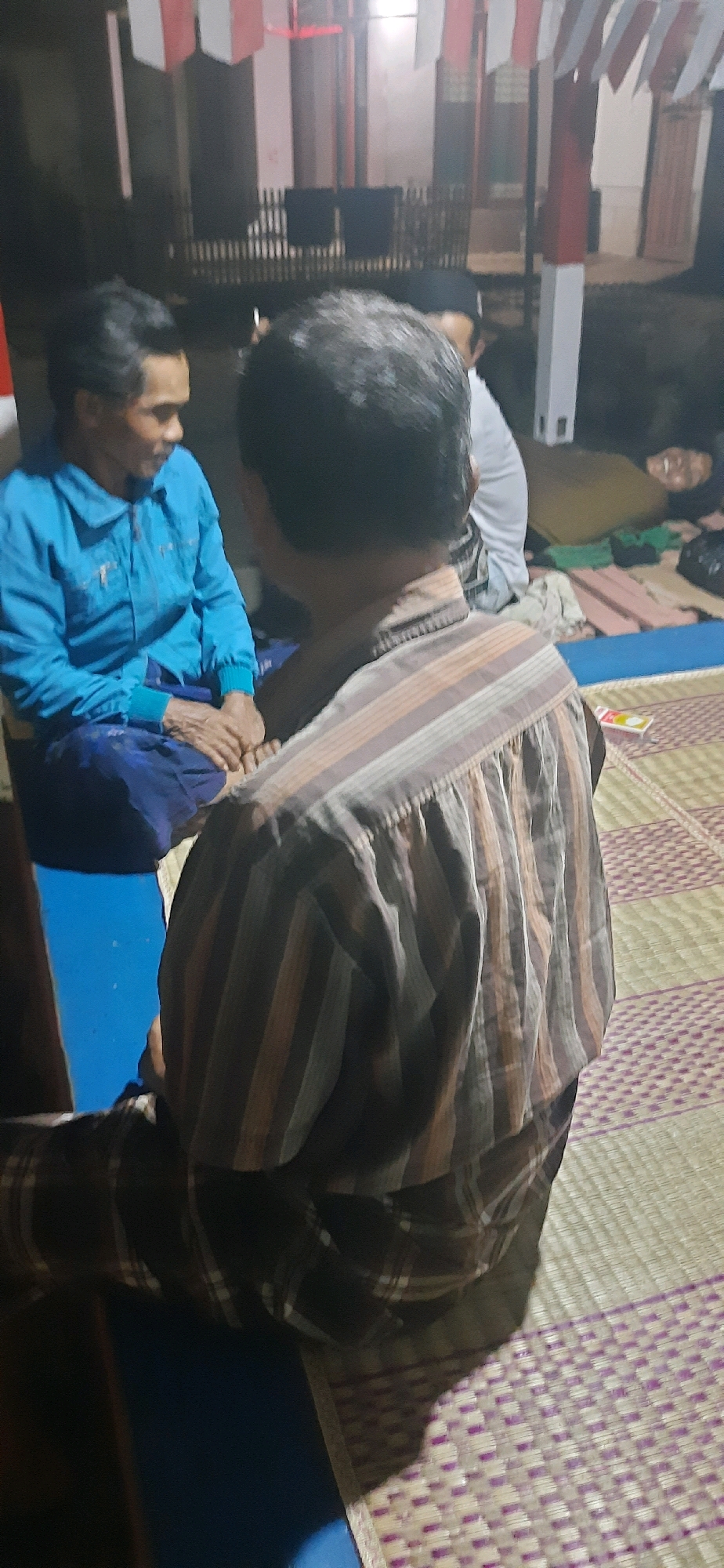In Desa Jayasari, Indonesia, two men are seated on a woven mat. The man on the left wears a bright blue jacket and blue pants, and is looking towards the man on the right. The man on the right, facing away from the camera, wears a striped collared shirt in shades of brown, orange, and white, and checkered pants. Behind them, a person in a white shirt is visible, and further in the background, there are red and white flags hanging, suggesting a festive or ceremonial occasion. The environment appears to be outdoors or in an open-air setting, possibly a yard or communal area, with a dimly lit background that shows structural elements like posts and a railing.