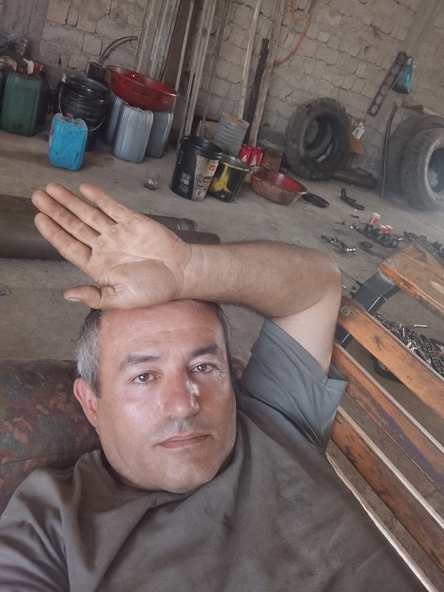 A man with graying hair and brown eyes, wearing a gray t-shirt, lies on a patterned surface, possibly a couch or chair. His left arm is raised, with his hand resting on his forehead. The background appears to be an interior space, likely a workshop or garage, with a brick wall and a concrete floor. Various objects are visible, including stacks of containers, buckets, tires, and what seem to be tools and spare parts scattered on the floor and on a wooden shelf unit. The lighting is dim, suggesting an indoor environment with limited natural light. The overall impression is of a relaxed or possibly fatigued individual in a work-related setting. The location is identified as Sabir Rakhimov, Uzbekistan.