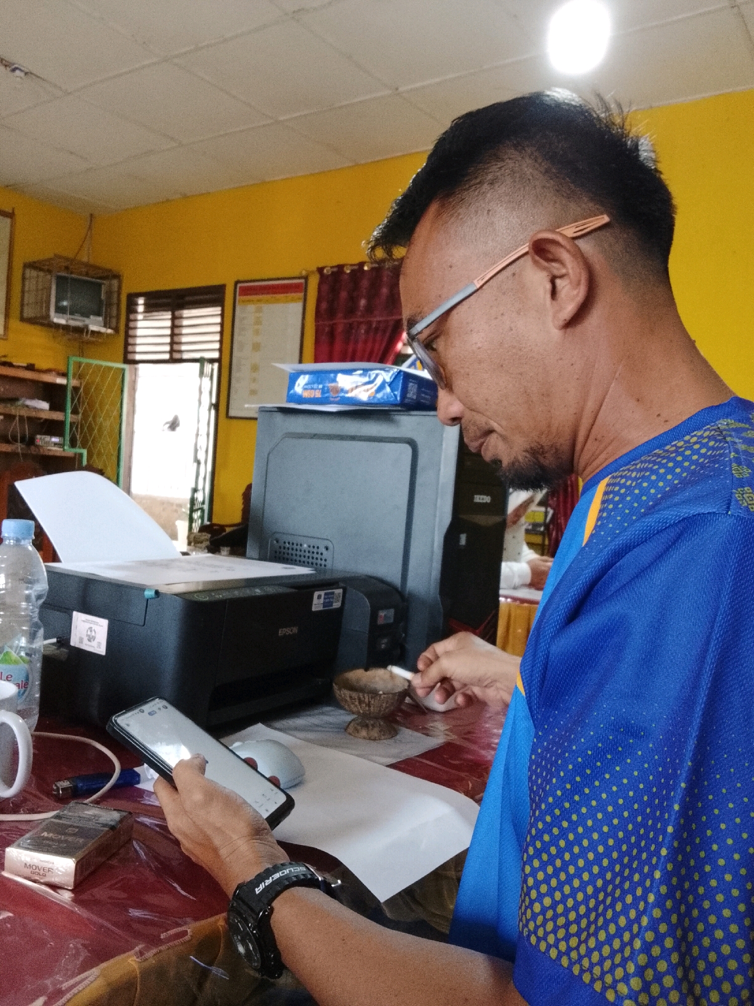 A man with short, dark hair and glasses is sitting at a table, holding a smartphone in his left hand and a cigarette in his right hand. He is wearing a blue t-shirt with yellow polka dots. On the table, there is a printer, a computer tower, a bottle of water, a cigarette pack, a lighter, and a small ashtray. The room is painted yellow and has a white ceiling with fluorescent lights. In the background, there is a window with blinds, a small television, and some shelves with papers and books. The scene appears to be an office or a home study.