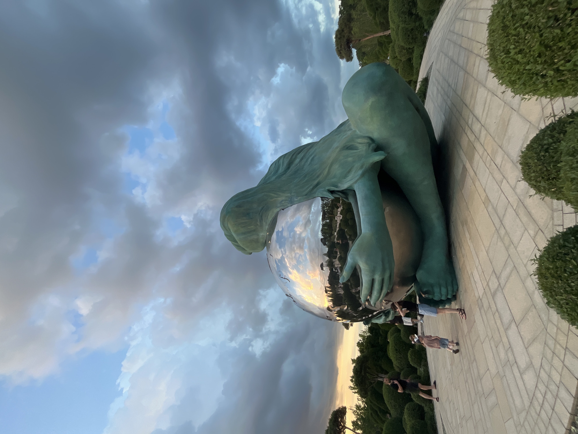 The image depicts the "World Child" sculpture, a large bronze statue of a child cradling a sphere, located in Krasnodar, Russia. The sculpture is situated on a paved plaza surrounded by meticulously trimmed bushes and trees, suggesting a park or garden setting. The sky above is dramatic, with heavy, dark clouds interspersed with patches of lighter blue and hints of sunset colors near the horizon. Reflections of the sky and surrounding landscape are visible on the polished surface of the sphere. Several people are present in the scene, including a man in a black shirt and shorts standing on the left, and a woman and a child in the foreground, also on the left, who appear to be observing the sculpture. Another person is visible in the background. The overall impression is of a contemporary art installation within a landscaped urban environment, under a turbulent sky.