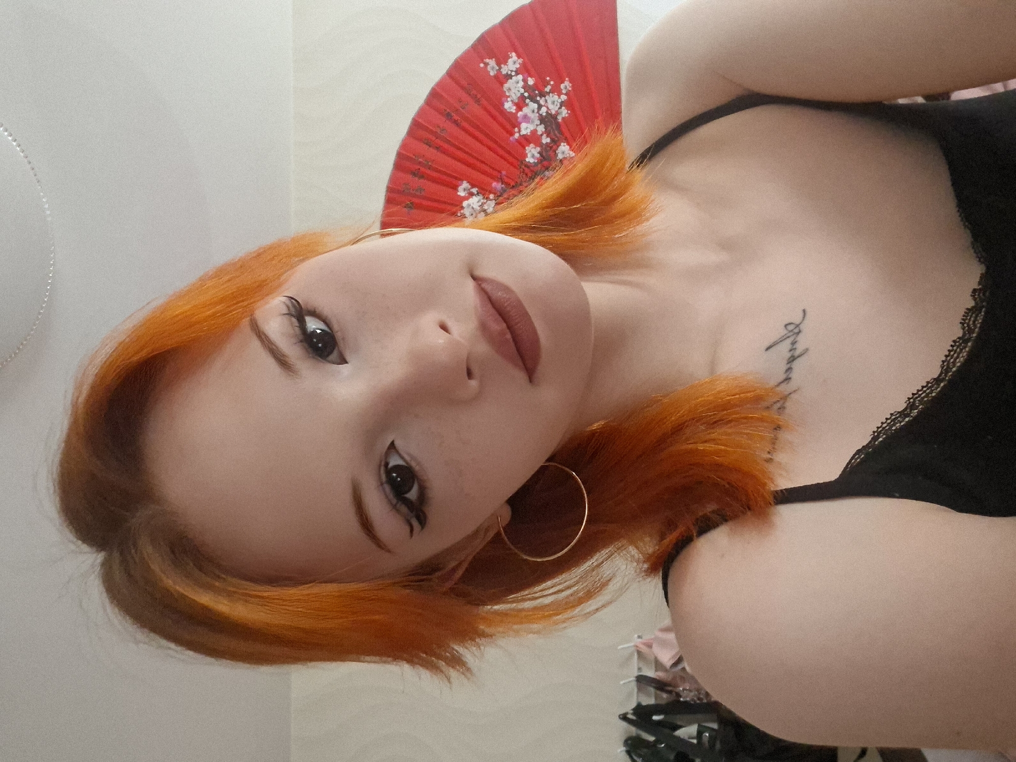 A woman with bright orange hair and fair skin is posing for a selfie indoors. She is wearing a black tank top with lace trim and large gold hoop earrings. The woman has visible winged eyeliner and a tattoo on her chest. A red folding fan decorated with white floral designs is behind her head. The setting appears to be a room in a house or apartment. There is no information to discern the exact location in Chaikovsky, Russia.