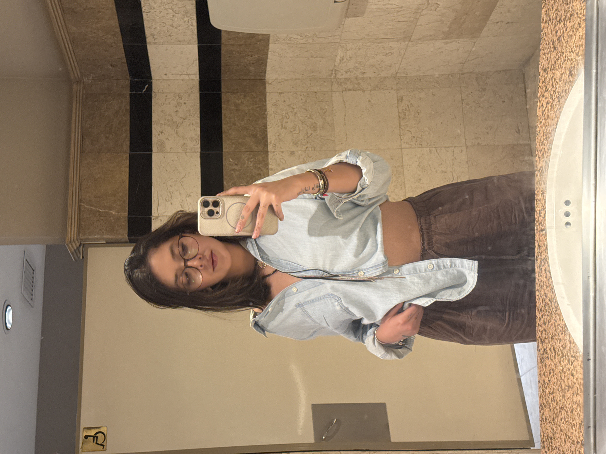 The image depicts a female individual taking a mirror selfie in what appears to be a public restroom in the city of Puebla, Mexico. The subject, centered in the frame, has shoulder-length dark hair and wears round-framed eyeglasses. She is clad in a light blue denim or chambray shirt, unbuttoned to reveal a portion of her midriff, and dark brown, loose-fitting pants. Her right hand holds a light-colored smartphone, featuring a multi-lens camera array, directed towards the mirror to capture the reflection. Her left hand is positioned near her waist, partially tucking into her waistband while grasping the bottom hem of her shirt. Accessories include multiple bracelets on her right wrist, a ring on her left hand, and a dark necklace visible around her collarbone. The restroom environment features a plain, light-colored wall to the left, which includes a metallic door handle and a yellow sign bearing a black wheelchair accessibility symbol. The wall to the right is covered in light beige tiles with two prominent horizontal dark brown or black stripes. Above these tiles, decorative molding is visible near the ceiling, which is light-colored and contains a recessed circular light fixture and a rectangular vent. A white dispenser-like object is mounted on the tiled wall. The foreground displays a speckled, light brown countertop with the curved edge of a white sink basin.