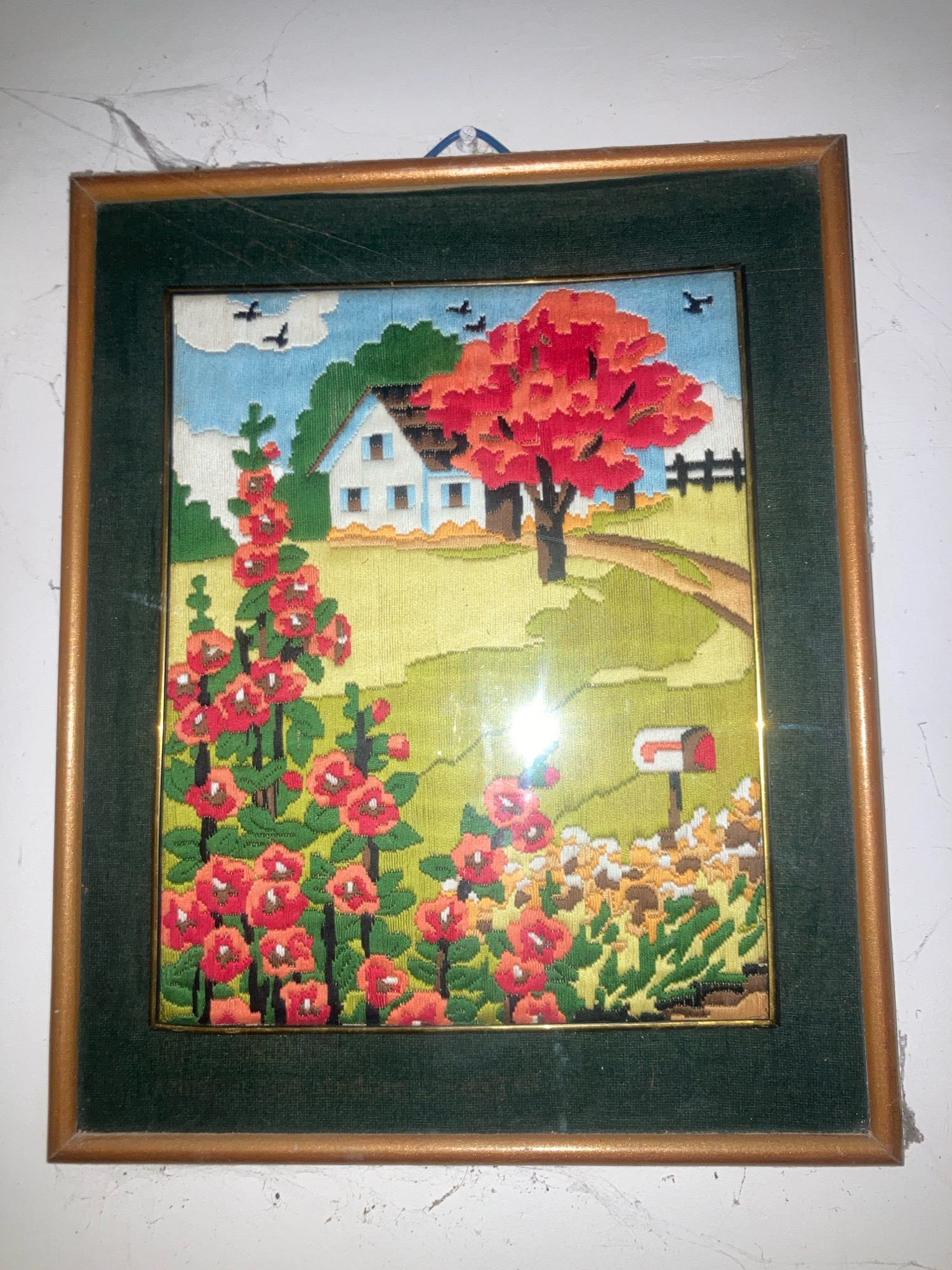 The image shows a framed needlepoint artwork depicting a rural landscape. The artwork features a white house with blue windows, a vibrant red-leafed tree, a dirt path, a mailbox, and a field of red and orange flowers, likely hollyhocks. Several black birds are depicted flying in the light blue sky. The artwork is framed with a dark green mat and a wooden frame. It is hung on a white wall that appears to have some cobwebs. The location of this artwork in Daladagama, Sri Lanka, is not discernible from the image itself.