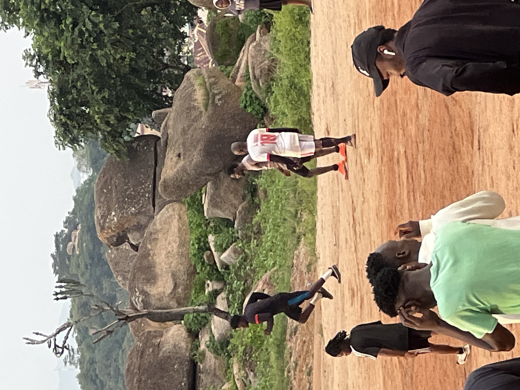 The image captures a dusty, outdoor scene with a group of young men engaged in physical activity, possibly a soccer practice or casual game, in Jos, Nigeria. The terrain is a reddish-brown dirt ground, characteristic of many sports fields in the region. In the background, large rock formations dominate the landscape, interspersed with lush green vegetation and trees, hinting at the hilly topography of Jos. A few buildings are visible in the distance, some with corrugated iron roofs.
Several individuals are the focus of the image. In the mid-ground, a person in a black athletic outfit with a blue stripe on the shorts is captured mid-stride, running from left to right. To their right, another person wearing a white t-shirt with the number "18" in red, and white shorts, appears to be walking or jogging. Further to the right, a person in a grey hoodie and black shorts stands observing. In the foreground, partially visible, are individuals in casual attire. One person wears a light green t-shirt and is looking down, with their hands near their head. Another person in a black jacket and baseball cap with visible earbuds is looking to the right. A person in a black and white athletic outfit is also visible to the left, facing away from the camera. The lighting suggests it is either early morning or late afternoon, with the sky appearing overcast. The overall impression is of a lively, communal activity taking place in a natural, rugged environment.