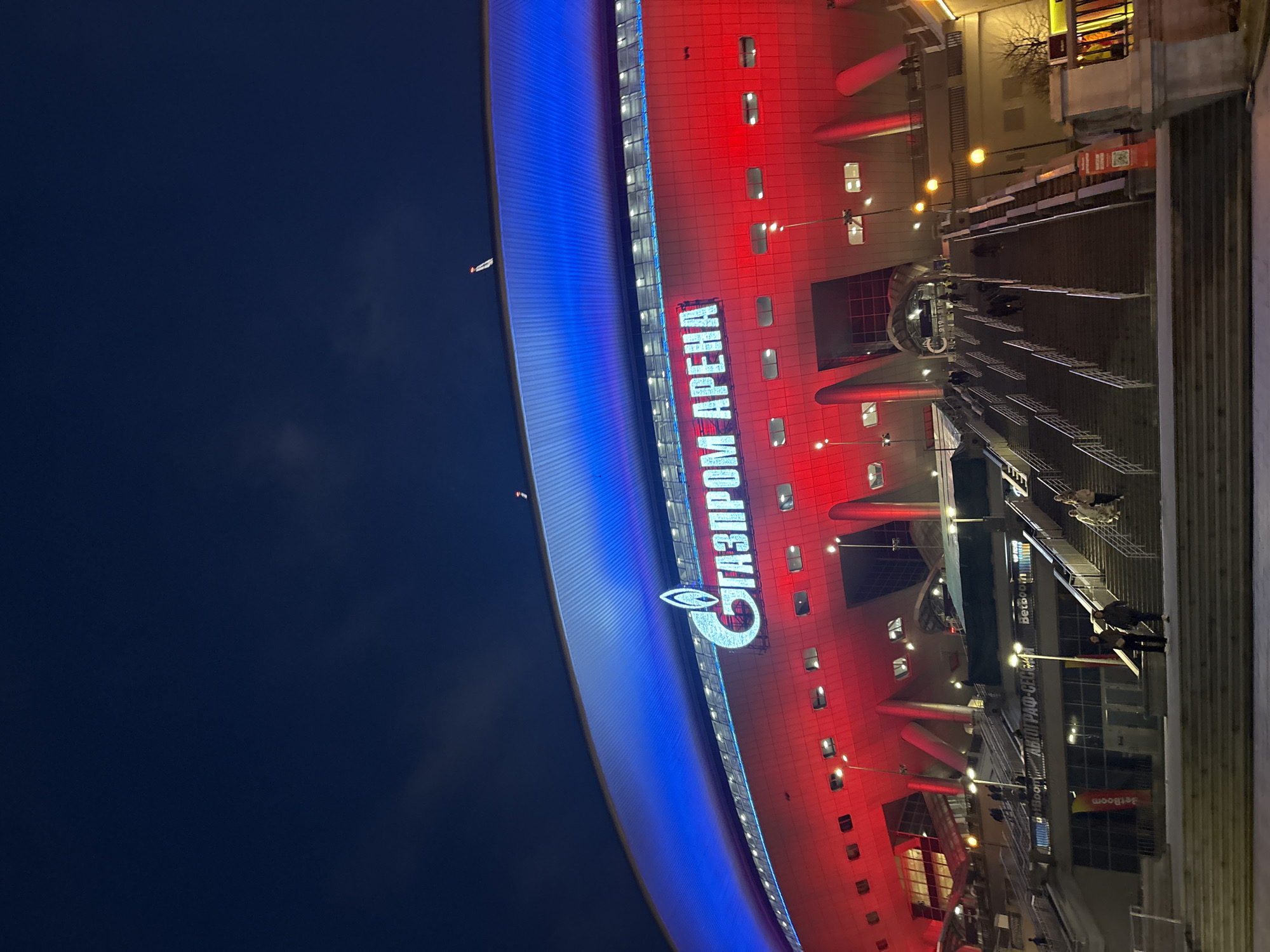 Captured from a low-angle perspective at night, the image features the Gazprom Arena in Saint Petersburg, Russia. The stadium's exterior is illuminated in blue and red, with the "Газпром Арена" (Gazprom Arena) signage prominently displayed. The surroundings consist of stairs and walkways leading up to the stadium's entrance. A few individuals can be seen walking up and down the stairs. Streetlights illuminate the pathways, and some trees are discernible in the background. The night sky is visible above the stadium.