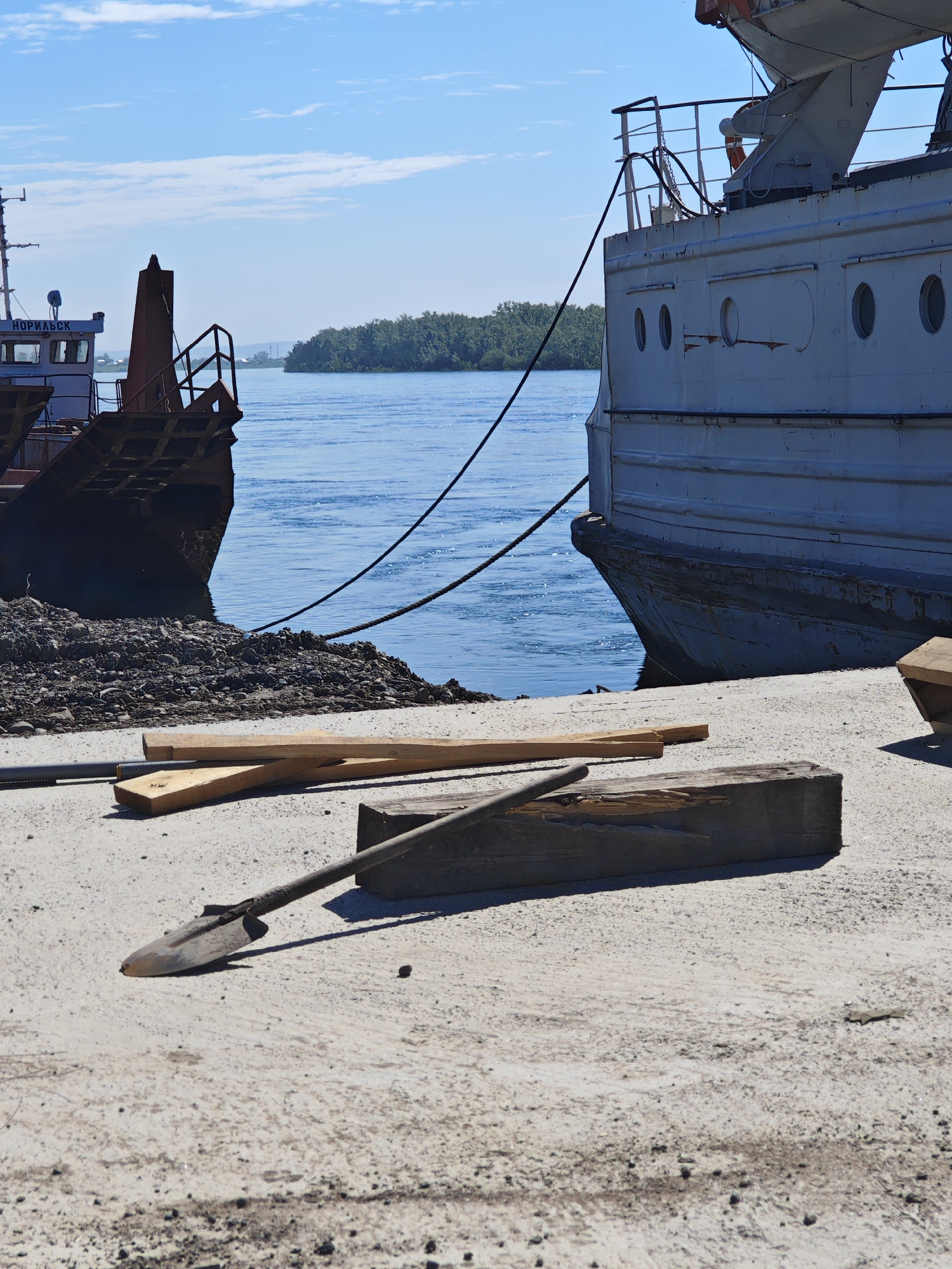 The image depicts a waterfront scene, likely in the city of Krasnoyarsk, Russia, given the Cyrillic text "НОРИЛЬСК" visible on the side of a vessel. In the foreground, a weathered concrete pier is visible, with several wooden planks and a shovel lying on its surface. The shovel is positioned diagonally, with its blade pointing towards the left and resting on a piece of lumber. Beyond the pier, two ships are docked. The vessel on the left appears to be a cargo ship or ferry, with a visible section of its deck and railings. The vessel on the right is larger, with a gray hull and several circular windows, tied to the pier with thick ropes. The water is calm and blue, reflecting the clear sky. In the background, a treeline of lush green foliage borders the river or bay, suggesting a natural landscape. The overall impression is one of a working port, with elements of both industrial activity and natural beauty.