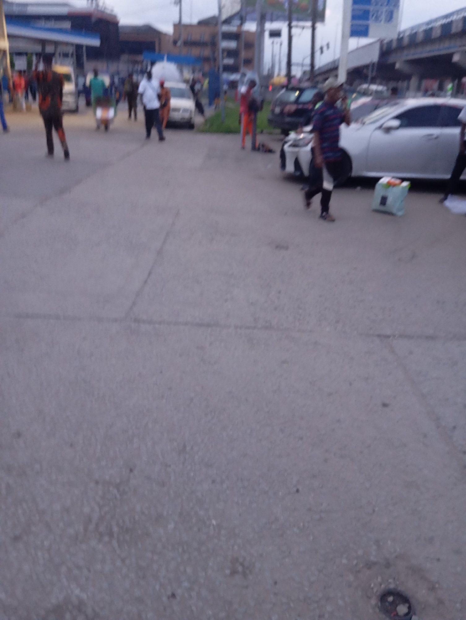 The image depicts a street scene in Port Harcourt, Nigeria, likely in the evening or overcast conditions given the lighting. Several individuals are visible on a paved road. In the foreground, a person in a striped shirt and dark pants walks towards the right, holding a phone to their ear. To their left, a light blue bag with an orange handle sits on the ground. Further in the background, a person in a patterned outfit walks from left to right, followed by another person in a white shirt and dark pants. A white car is parked on the right side of the road. In the mid-ground, to the left, a person in a green shirt stands near a vehicle, and a cart with a rider is visible. The background shows buildings and overpasses, typical of an urban environment. The overall scene suggests a bustling public space with pedestrian and vehicular traffic.