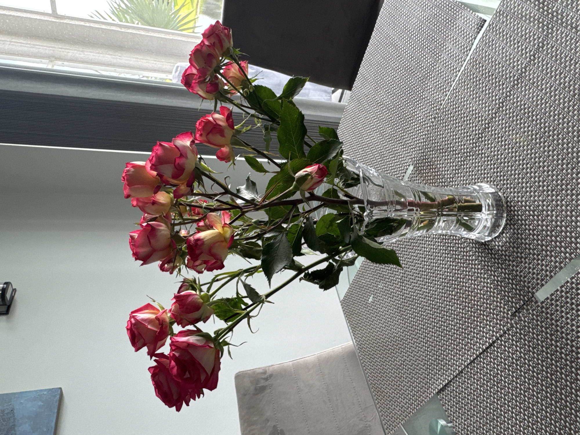 A bouquet of pink and white roses is arranged in a clear glass vase on a dining table. The table is covered with a textured placemat. Two chairs are visible, one on the left and one on the right side of the table. In the background, a window with curtains allows natural light to enter the room, and a piece of artwork hangs on the wall. The overall setting appears to be a residential dining area.