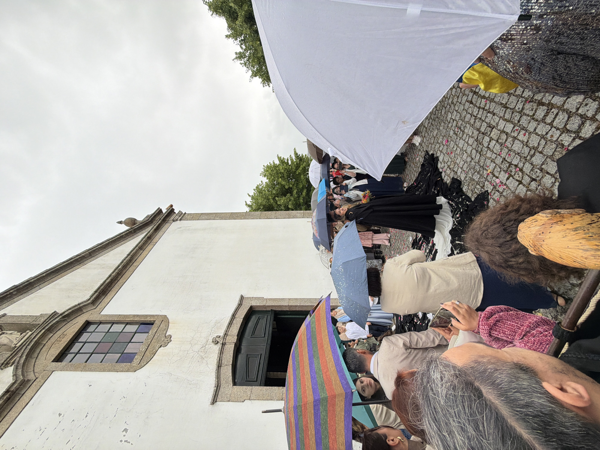 The image captures a gathering outside a white building with a dark green door and a stained glass window, likely a church, in Vila Real, Portugal. People are holding umbrellas of various colors (white, striped, blue) suggesting it is raining. Some are dressed formally in black and white, possibly for a wedding. There are trees visible in the background and cobblestones on the ground. The scene appears to be at a celebration after an event.