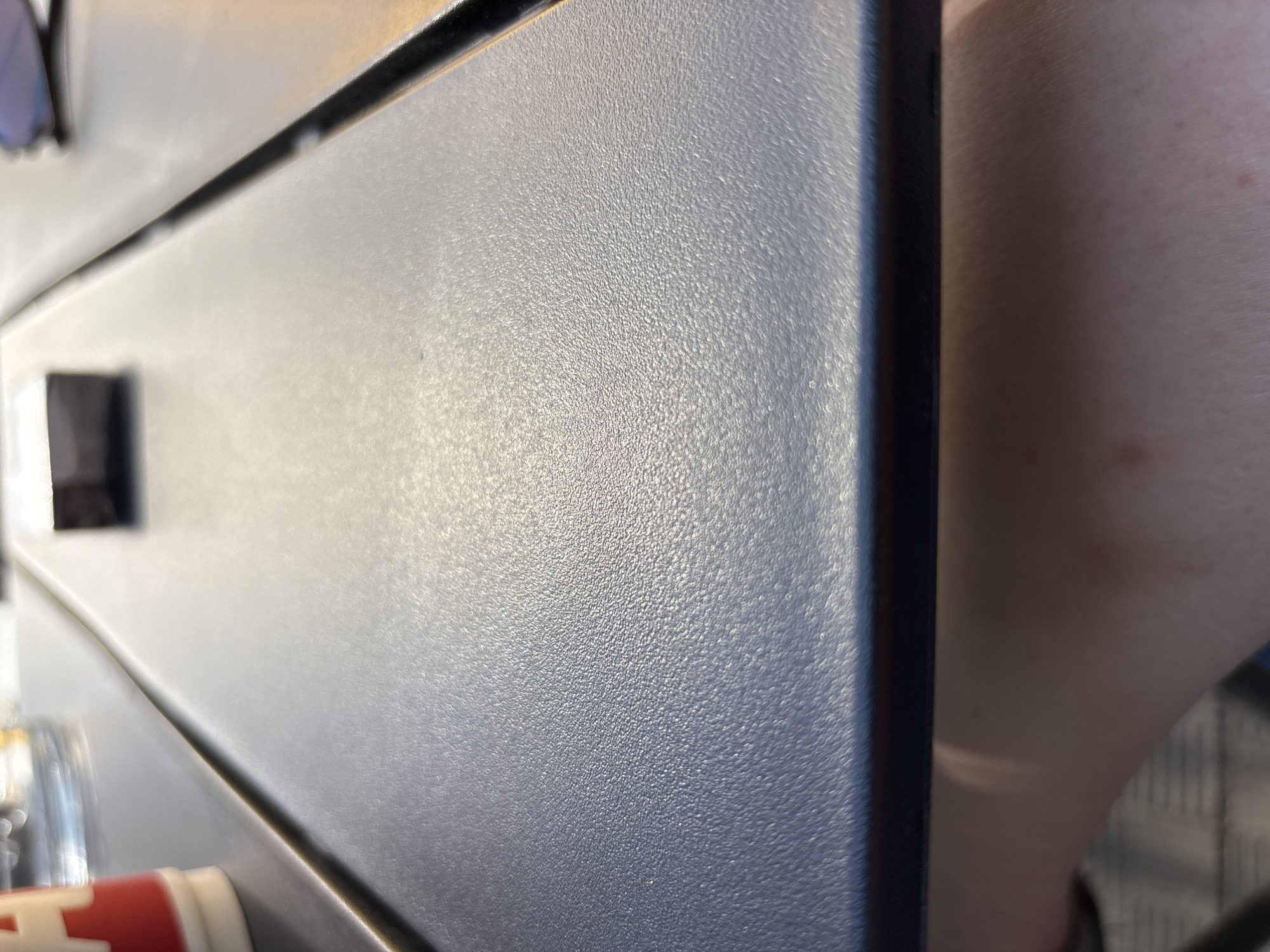 The image displays a close-up, low-angle view of a dark, textured surface, likely a table, in an indoor setting. The surface is illuminated by bright, directional light, creating strong highlights and shadows. A portion of a red and white cup is visible in the upper left corner, partially out of focus. In the upper center, a small, dark, rectangular object, possibly a pen or a small electronic device, rests on the surface. The bottom portion of the image shows a person's arm and skin, indicating the viewpoint is from someone seated or leaning at the table. The environment appears to be a public space such as a cafe or restaurant, but no identifying features of the location are discernible. The lighting suggests it could be daytime, with natural light filtering in.