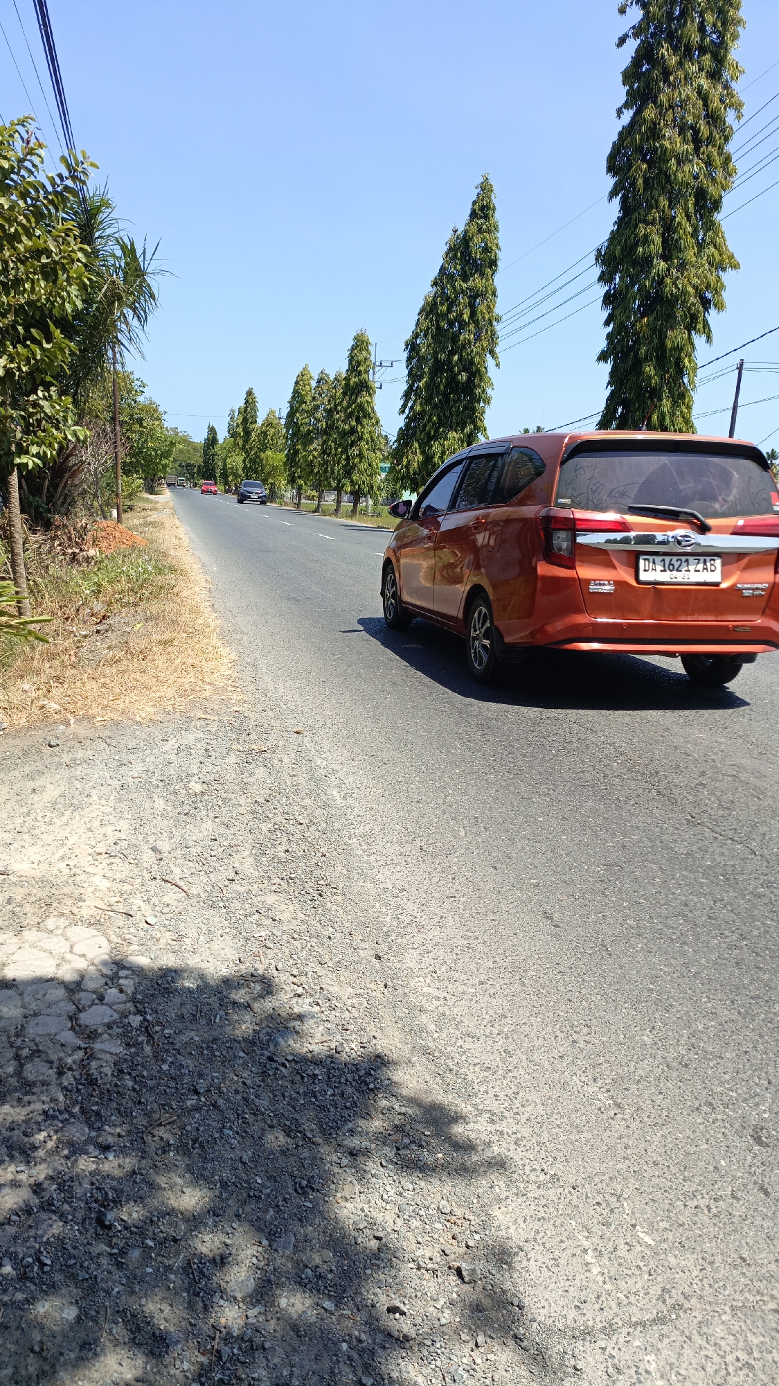 The image depicts a rural road in Mudalang, Indonesia, under a clear blue sky. The road is paved with asphalt and has a gravel shoulder on the left side. Tall, slender trees line the right side of the road, interspersed with power lines. On the left side, there are more trees and some dry vegetation. A bright orange car, identified by its license plate DA 1621 ZAB, is in motion, heading away from the camera. Further down the road, a dark-colored car and a red car are also visible, suggesting light traffic. Shadows from trees are cast across the road and shoulder, indicating a sunny day. The overall scene is one of a typical Indonesian road in a semi-rural area.