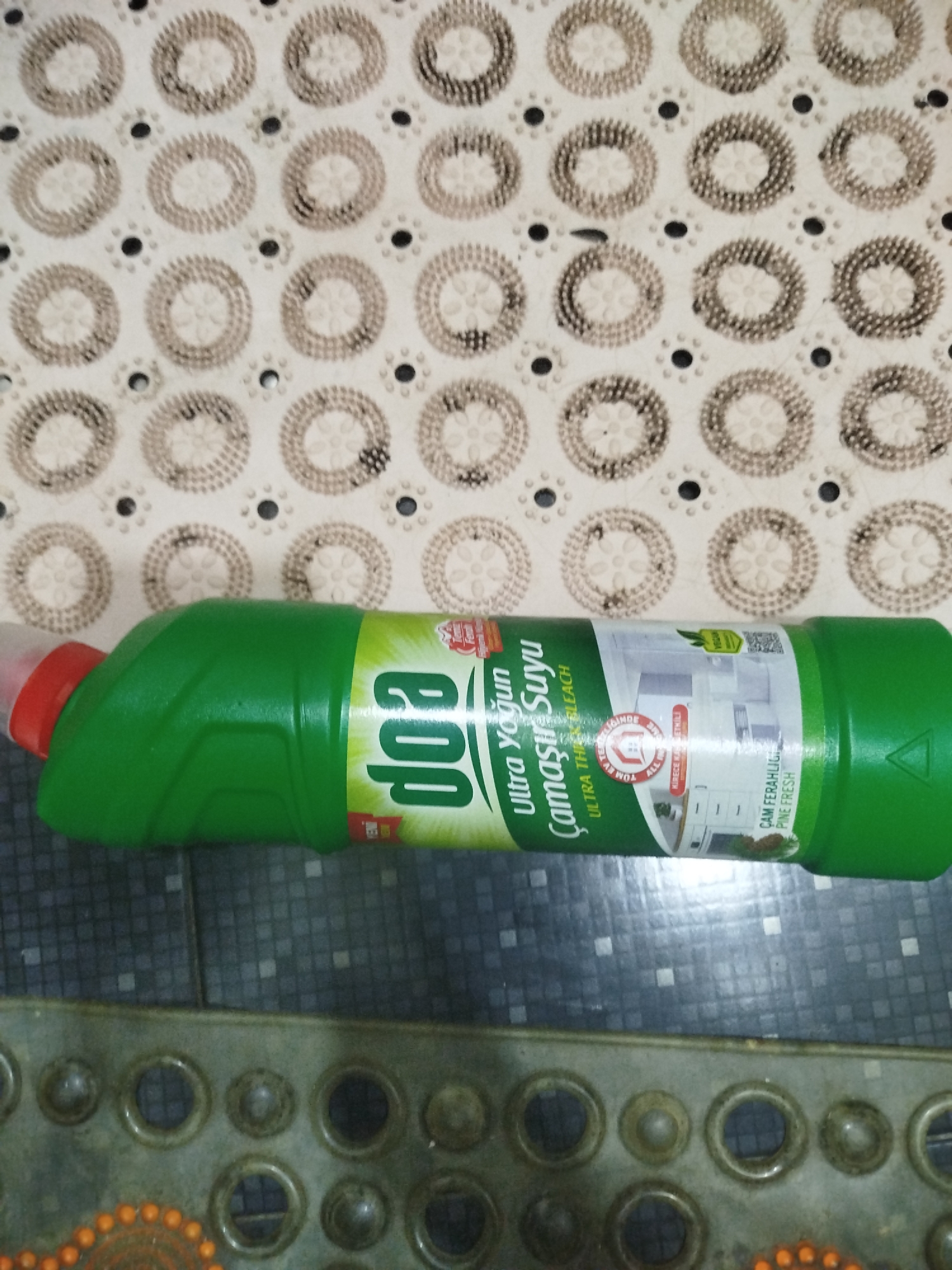 The image shows a green plastic bottle of "Dop Ultra Yoğun Çamaşır Suyu" (Dop Ultra Concentrated Laundry Bleach) with a red cap. The bottle is lying horizontally on a dark, tiled surface, with a textured, light-colored mat with circular patterns visible in the background. A portion of a gray, circular-patterned mat or object is in the foreground. The scene appears to be indoors, possibly in a bathroom or laundry area. There are no people or animals present. The specific location within Batumi, Georgia, cannot be determined from the image.