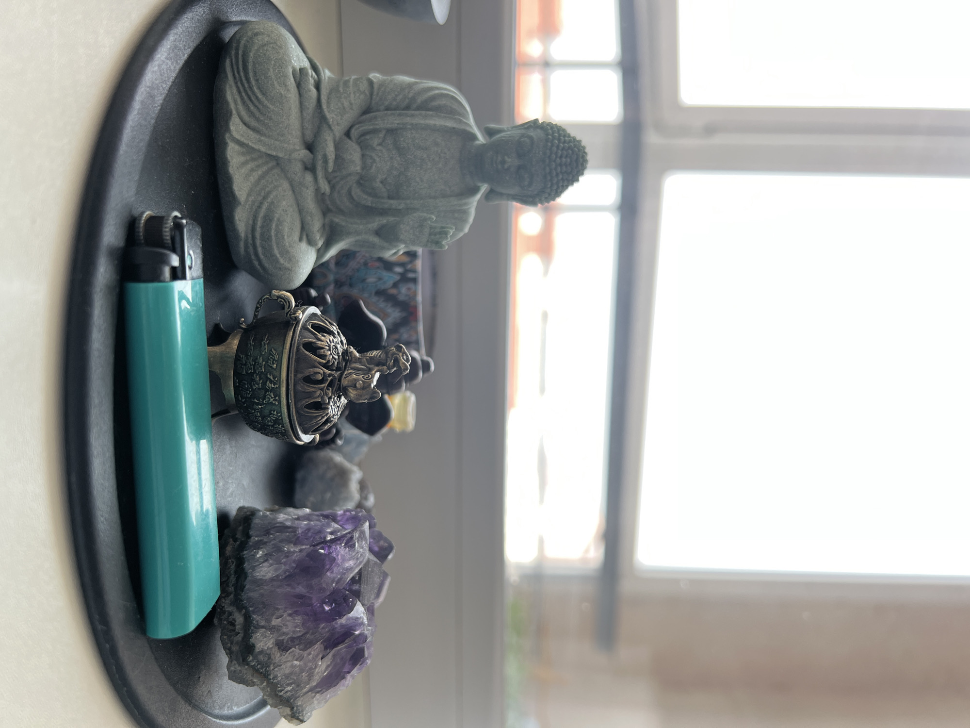 The image depicts a still life arrangement on a black circular tray placed on a white surface, likely a windowsill or table. Positioned on the tray are several items: a small, grey, seated Buddha statue with its right hand raised in a gesture of blessing or protection, an ornate bronze incense burner with a lid featuring animalistic figurines, a cluster of raw purple amethyst crystals, and a teal-colored lighter lying horizontally in the foreground. Behind the objects, a window with white mullions and bright, blown-out daylight suggests an indoor setting, possibly during the daytime. The background is softly blurred, showing hints of indistinct shapes and colors. There are no people or discernable actions in the image. No specific location in Novosibirsk, Russia, can be identified from the visual information provided.