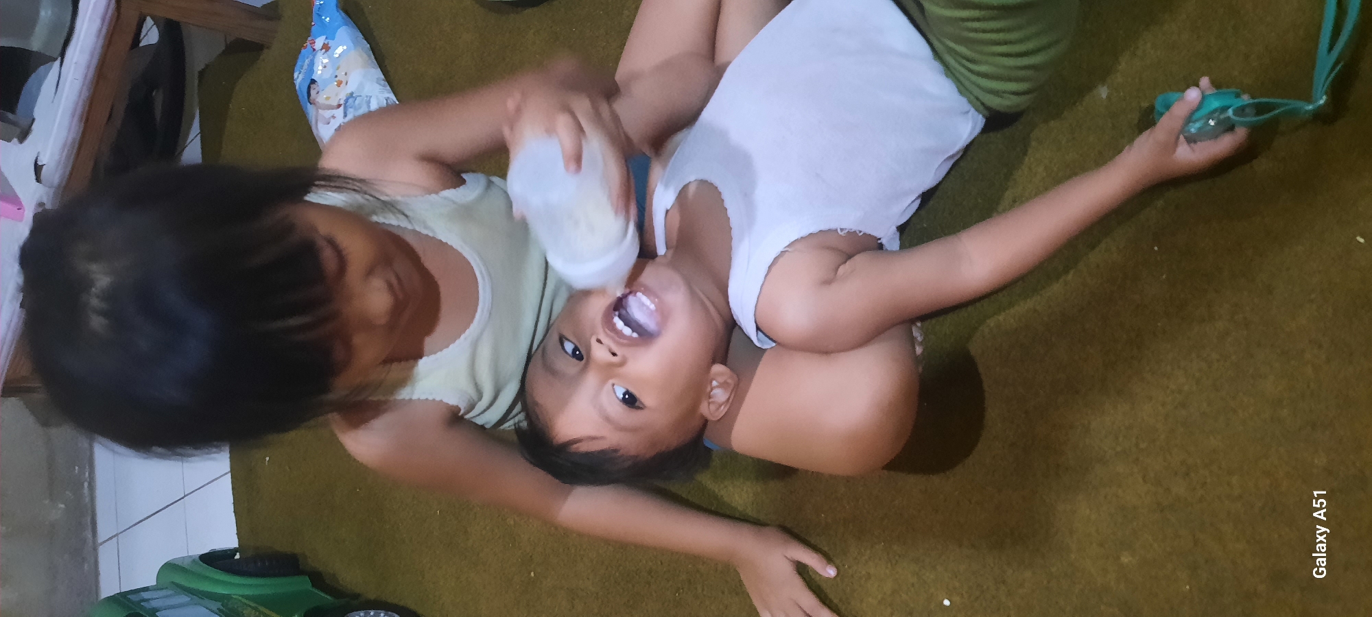 Two young children are on a green carpeted floor in a room in Jatitengah, Indonesia. The older child, with dark hair and wearing a light yellow tank top, is sitting and holding a baby bottle with milk. The younger child, lying on their back with their head in the lap of the older child, is wearing a white tank top and green striped pants. The younger child has their mouth open, and the older child is feeding them from the bottle. In the background, a toy green truck and a bag of snacks are visible. The overall scene depicts a moment of sibling care and interaction in a domestic setting.