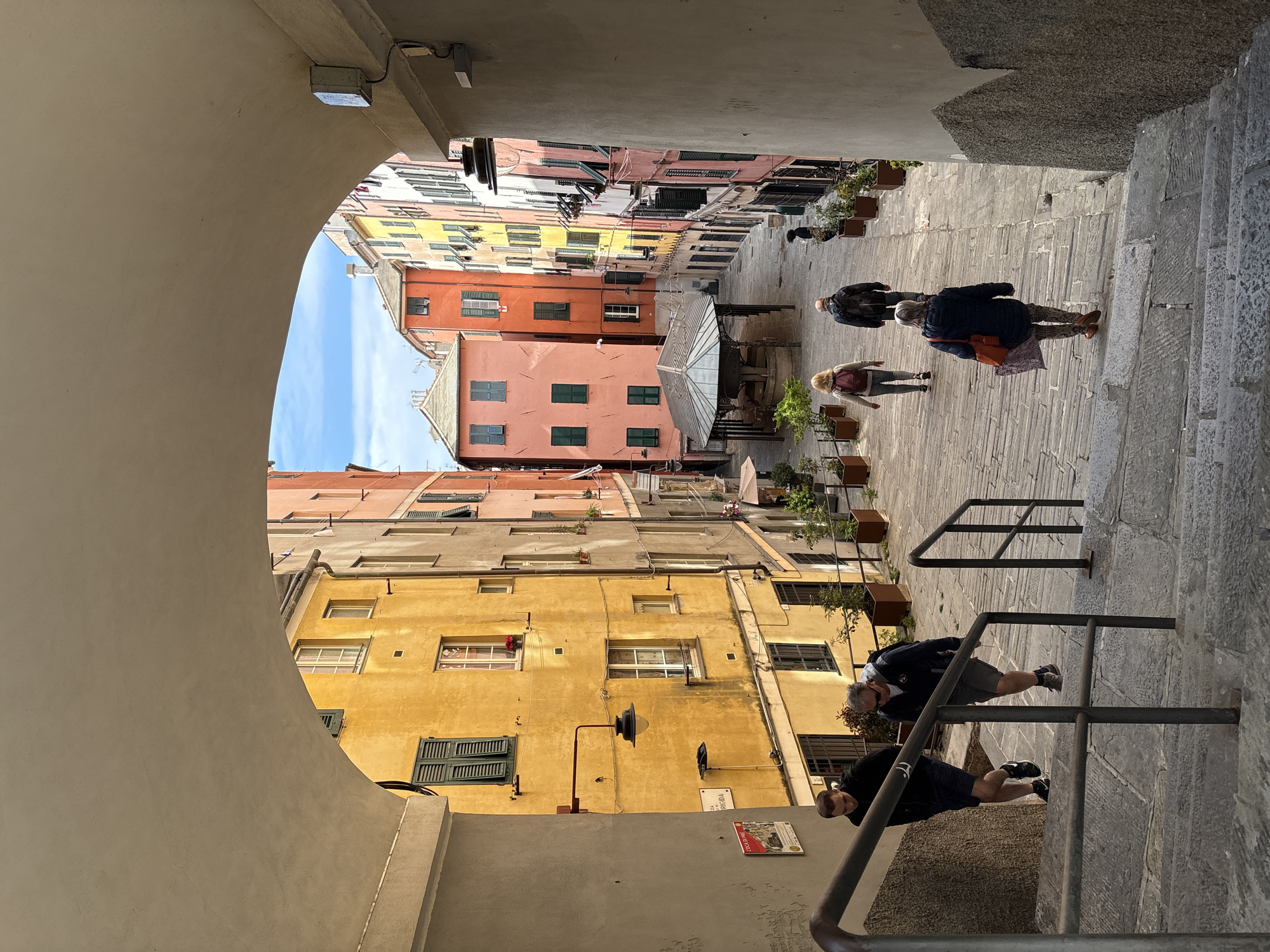 This image captures a vibrant, narrow street scene in Genoa, Italy, viewed from under an archway or covered passage. The archway frames the left side of the composition, extending into the foreground, and leads into a cobbled street paved with irregular stone slabs that gently descends towards the right.
Multi-story buildings with colorful, weathered facades, including shades of yellow, pink, orange, and reddish-brown, line both sides of the street. These buildings feature numerous windows, many adorned with dark green or brown shutters, some open, some closed. Planters with small green plants are strategically placed along the street and against the building walls, adding touches of greenery.
Several people are present, indicating a typical day. On the right side of the street, a set of stone steps with sturdy metal railings leads upwards. Two men are visible on these steps; one, closer to the viewer, wears a dark jacket and shorts, while another stands further up. On the cobblestone street below, a woman with blonde hair and a backpack walks away from the viewer. Another woman, dressed in a dark jacket with patterned pants and carrying a reddish-orange bag, walks towards the viewer. Additional pedestrians can be seen further down the street.
The sky, visible through a gap between the buildings in the mid-ground, is bright blue with some light clouds, suggesting a clear and sunny day, likely late morning or early afternoon given the shadows cast. A small structure with a glass roof, possibly a kiosk or shelter, stands on the street in the mid-distance. Utility wires and wall-mounted boxes are visible on the archway and buildings.
Visible text includes a white rectangular street sign on a yellow building, reading "VIA DI SCALA" and "VIORGIGIA" (likely "Via Orgigia"). Below this, a red sign with white text partially reads "DOLCE PIÙ". The scene conveys a sense of daily life in a historic and charming European urban environment.