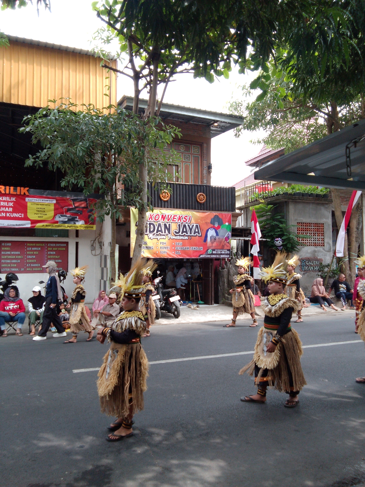 In Trenggalek, Indonesia, a cultural procession is underway on a city street. Several young boys, adorned in traditional attire consisting of feathered headpieces, straw skirts, and decorative armbands, are marching in formation. They are accompanied by onlookers seated on the side of the street and standing near buildings. Behind the procession, buildings with signage in Indonesian suggest local businesses, including a shop for "KRILIK" and another for "KONVEKSI." Red and white Indonesian flags are displayed, indicating a national celebration. The street is paved and lined with trees, with a cloudy sky overhead.