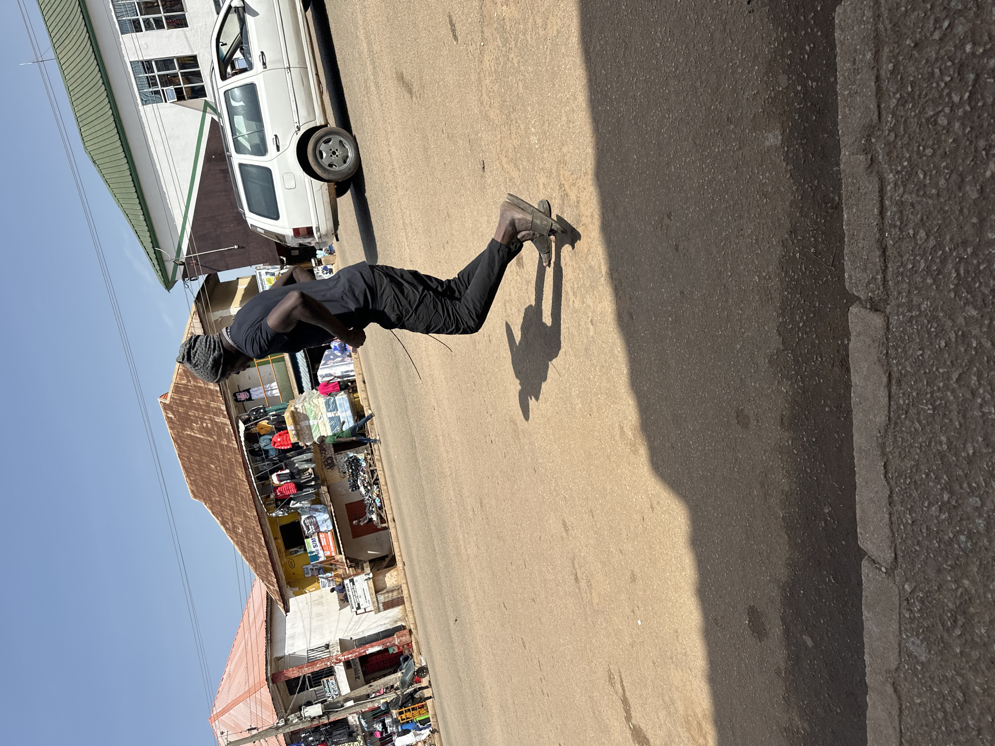 A street scene in Bukuru, Nigeria, features a young Black man in mid-stride, running across a dusty, paved road. He is wearing a black t-shirt, black pants, and dark sandals. His body is angled to the right, with his arms bent and in motion, indicating he is running. Behind him, a white station wagon is parked on the right side of the road, and several buildings line the left side of the street, some appearing to be shops with merchandise displayed outside. The sky is clear and blue, suggesting a bright, sunny day. The overall environment appears to be a bustling urban area with typical street activity.
