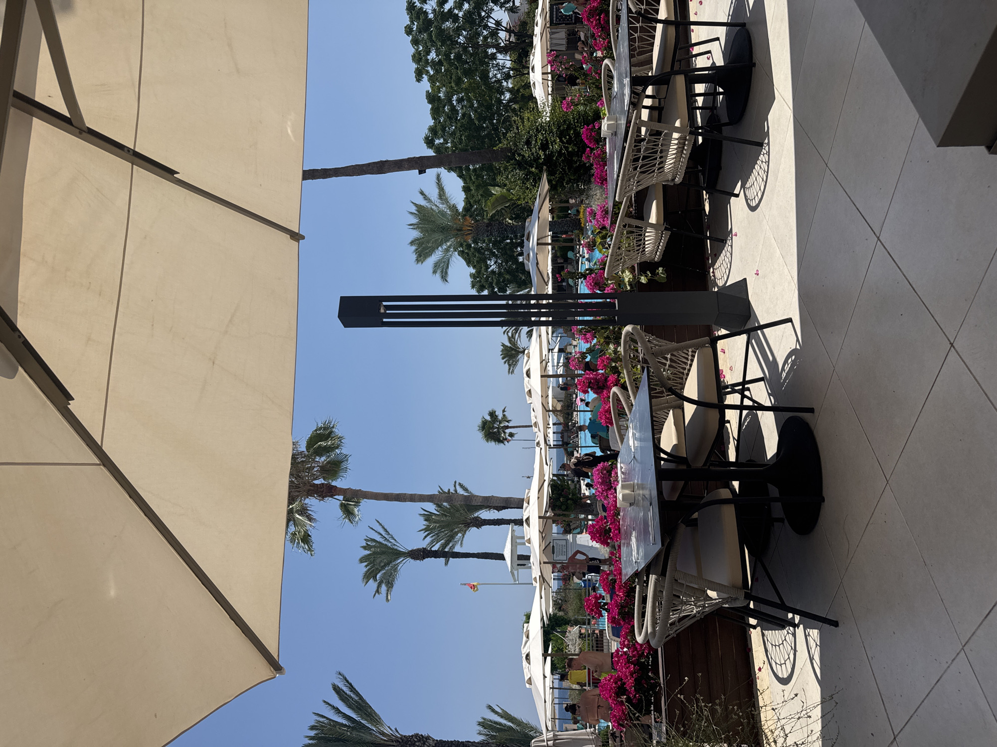 The image depicts an outdoor seating area at a resort in Kemer, Turkey. In the foreground, a black table with four chairs made of woven material and beige cushions is set up on a tiled patio. The table is set with white cups and what appears to be a menu. Behind the table, a low wall is adorned with vibrant pink flowers, with a tall black lamp post to the left. In the middle ground, several white umbrellas provide shade over more tables and chairs, with people visible lounging and walking around. A swimming pool with bright blue water is in the background, surrounded by lush green trees and palm trees. The sky is clear and blue, indicating a sunny day. The overall scene suggests a resort setting by the sea, common in Kemer.
