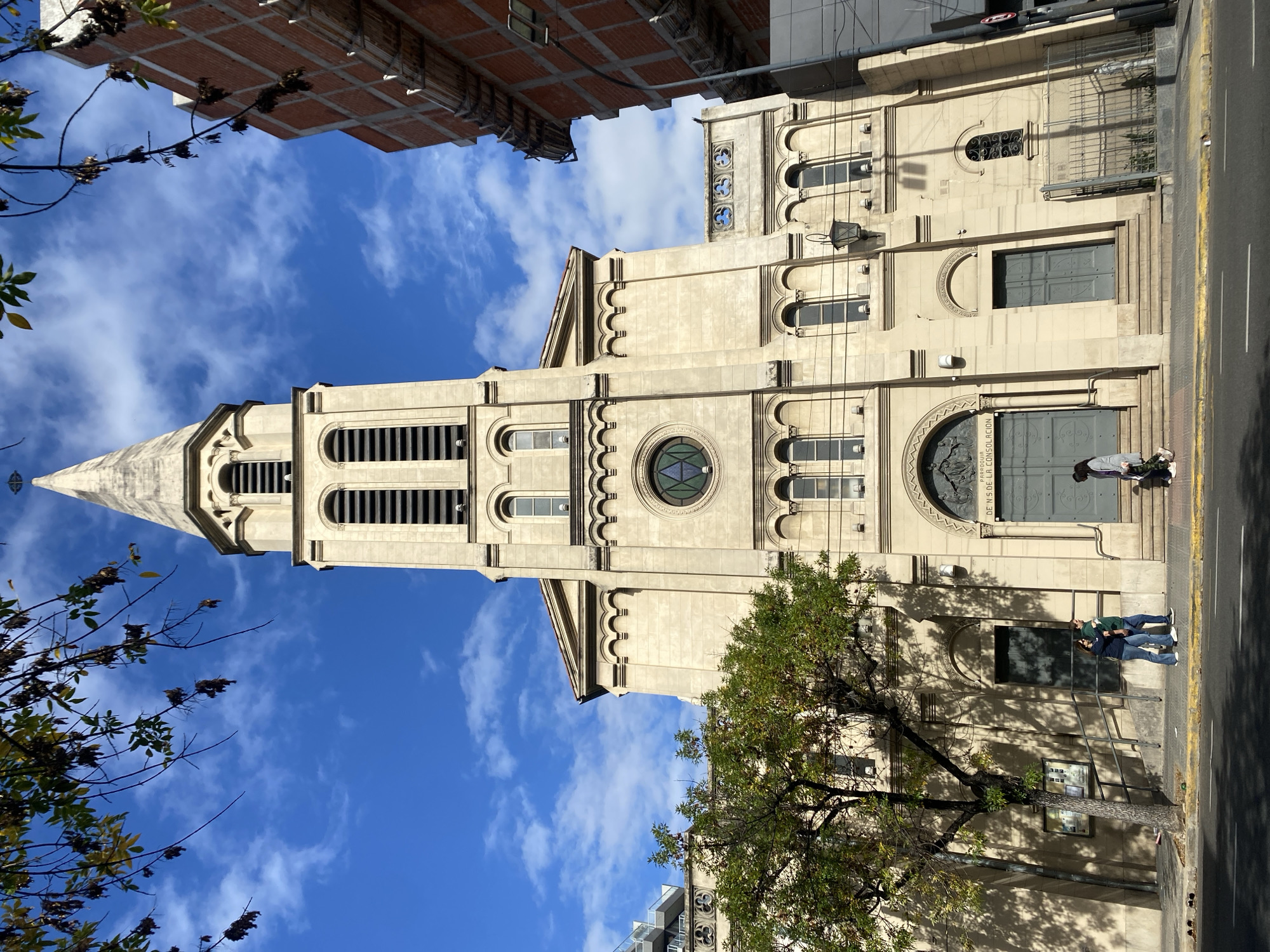 The image captures the Parroquia Nuestra Señora de la Consolación church in Buenos Aires, Argentina. The church is constructed from beige stone with a tall, pointed spire and several arched windows and doorways. A circular stained-glass window is also visible. The sky is blue with scattered white clouds. A portion of a brick building appears at the top left of the frame, while a tree with green leaves is positioned in front of the church on the right side. A couple is visible standing near the entrance of the church. The view is from a low angle, emphasizing the height of the church's tower. The building appears to be on the corner of a road with the sidewalk on the right side of the frame.