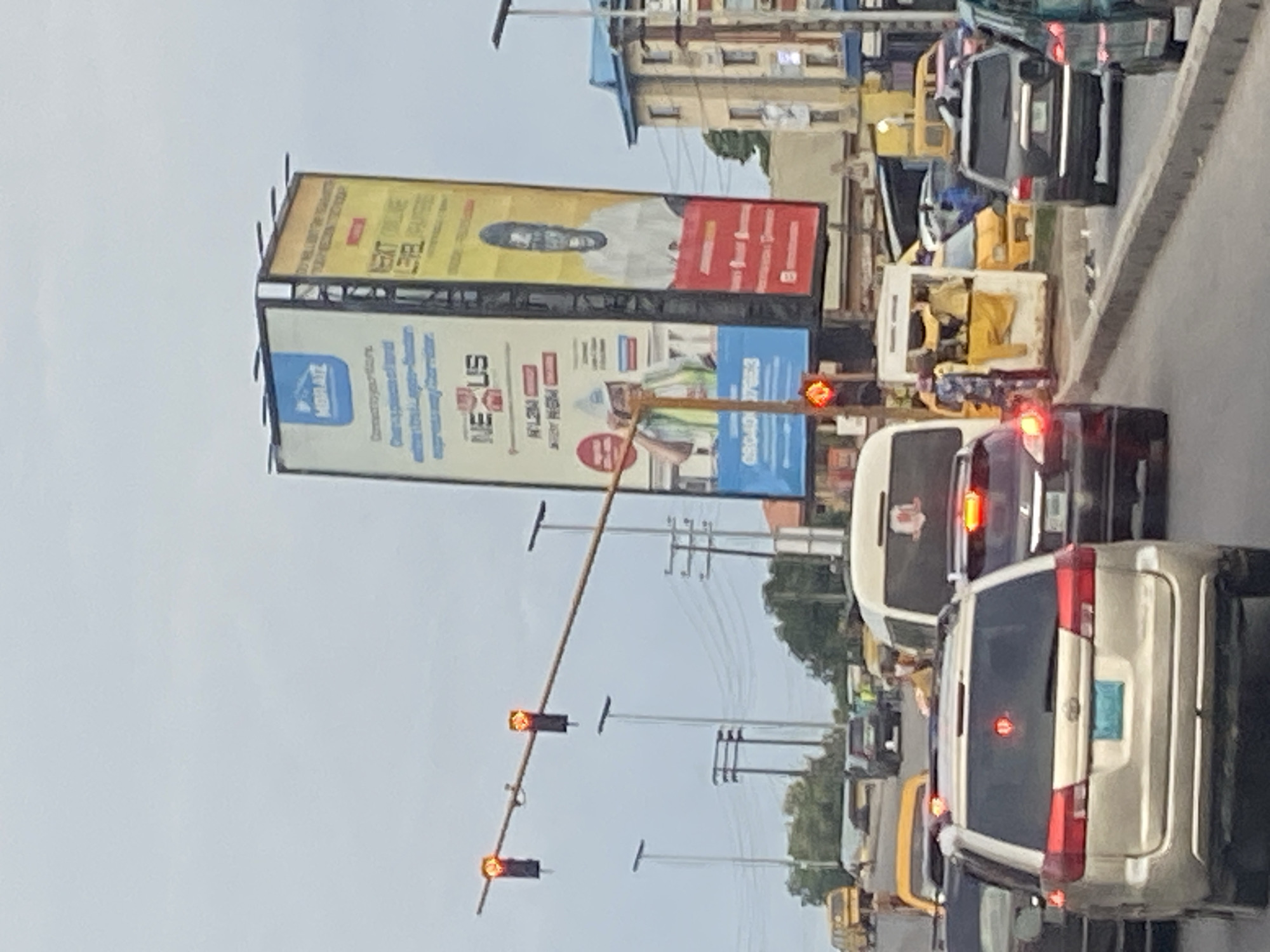 A street scene in Ikorodu, Nigeria, features a traffic jam on a cloudy day. Several cars, including SUVs and vans, are stationary on the road. Traffic lights are visible, with two red lights illuminated. Two large billboards stand prominently in the background. The left billboard advertises "MORAIZ" with a slogan about connecting to the Lagos-Ibadan expressway. The right billboard displays a portrait of a man and the text "NEXT LEVEL." In the background, buildings, trees, and utility poles line the street. A yellow minibus or taxi is parked to the right of the road, with people gathered around it.