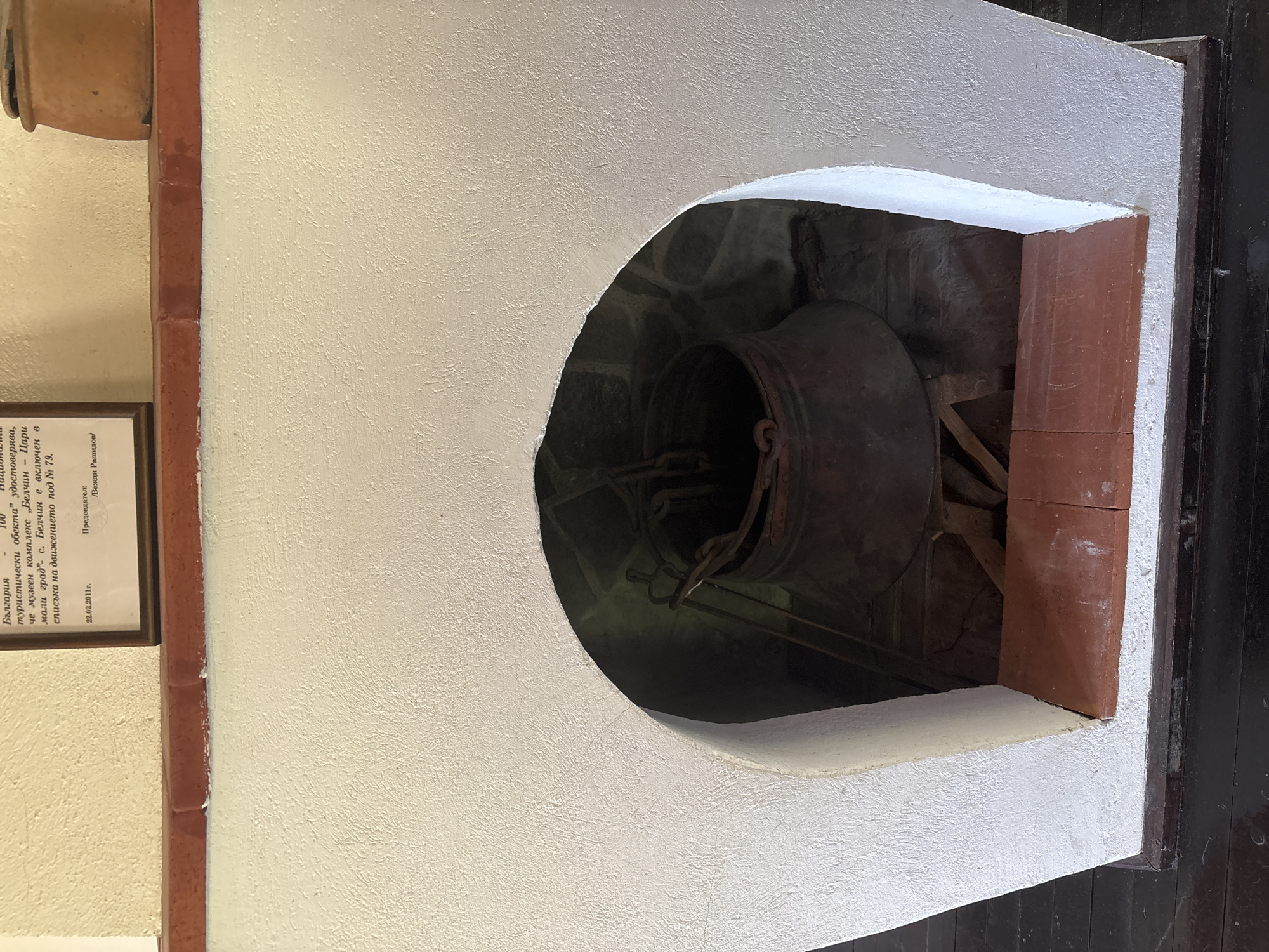 The image depicts an interior scene, likely within a historical or museum setting in Belchin, Bulgaria. In the foreground, a dark, worn wooden floor runs horizontally. Directly above it, a white, rough-textured, stucco-like structure forms the front of an arched alcove or hearth. A dark brown trim runs along the base of this white structure. The opening of the alcove is lined with two terracotta-colored tiles, forming a narrow, rectangular base.
Inside the dark, arched alcove, a large, dark, metallic pot or cauldron, possibly made of copper or bronze, is centrally positioned. This pot is supported by a rudimentary wooden stand with visible cross-braces. Dark metal chains descend into the alcove from an unseen upper attachment point, connecting to handles on the sides of the pot. The back wall of the alcove appears to be constructed of rough-hewn stones or masonry. The interior of the alcove is dimly lit.
Above the white alcove structure, a horizontal reddish-brown brick or tile border separates the white wall from an upper wall section. This upper section is painted a light beige or cream color and also exhibits a rough, textured finish. Mounted on this upper wall, slightly to the left of center, is a framed document or sign. The document contains text, partially visible in Cyrillic script, with a date "22.02.2011г." and names like "Вежди Рашидов", and references to "България" and "Белчин" as a "туристически обекта" (tourist object). To the far right of the frame, atop the reddish-brown border, a portion of a small, terracotta-colored pot or ceramic vessel is visible.
No individuals are present in the depicted scene. The lighting suggests an indoor environment, with illumination highlighting the frontal surfaces and diminishing into the depths of the alcove.