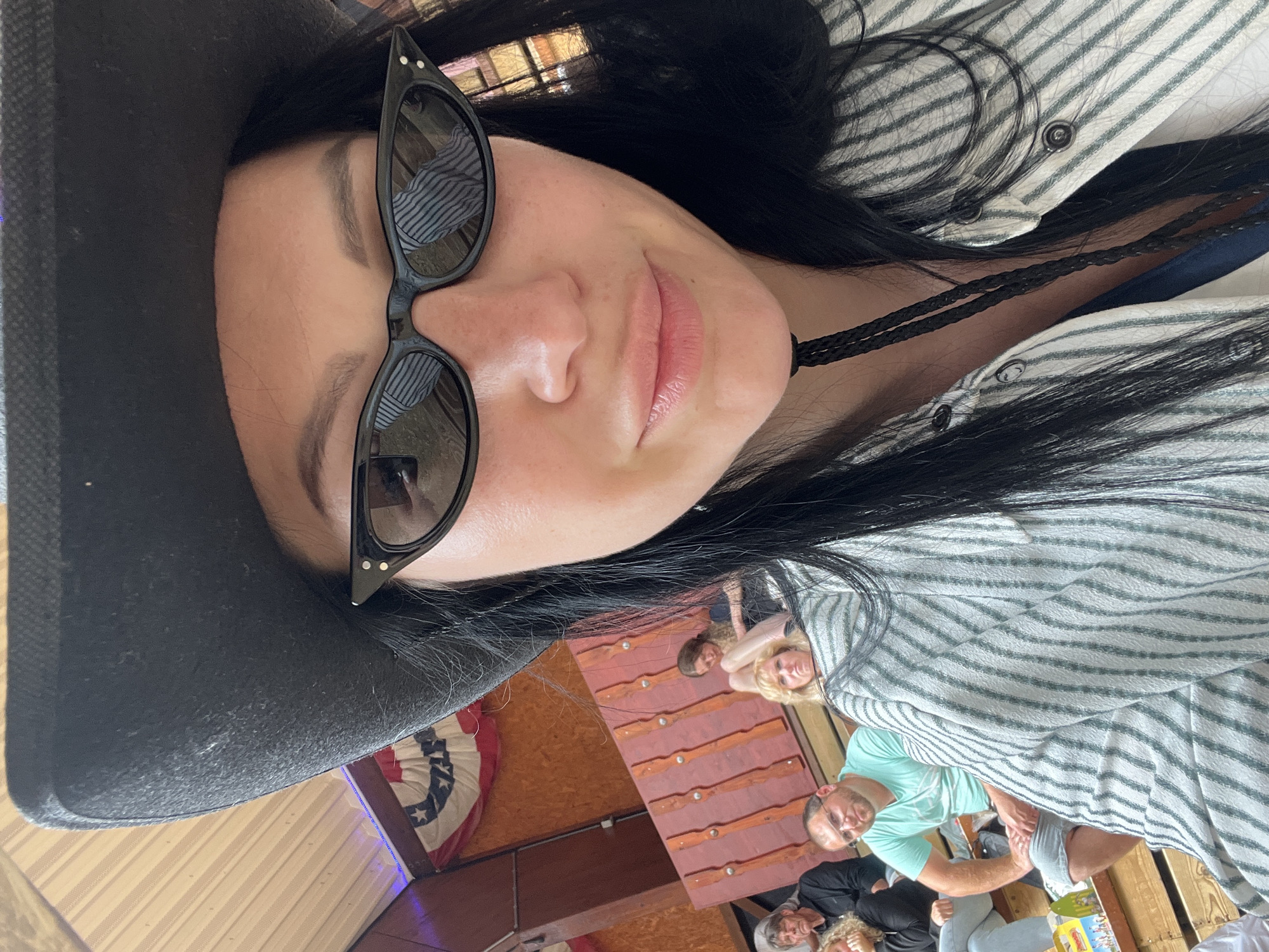 A close-up, selfie-style shot shows a woman wearing a black cowboy hat and black cat-eye sunglasses. Her dark hair is visible around her face. She is wearing a striped button-up shirt. In the background, out of focus, are several other people sitting on wooden benches. The environment appears to be an indoor venue with wooden paneling and a large American flag decoration. The lighting suggests it is daytime. The scene is likely a social gathering or event.