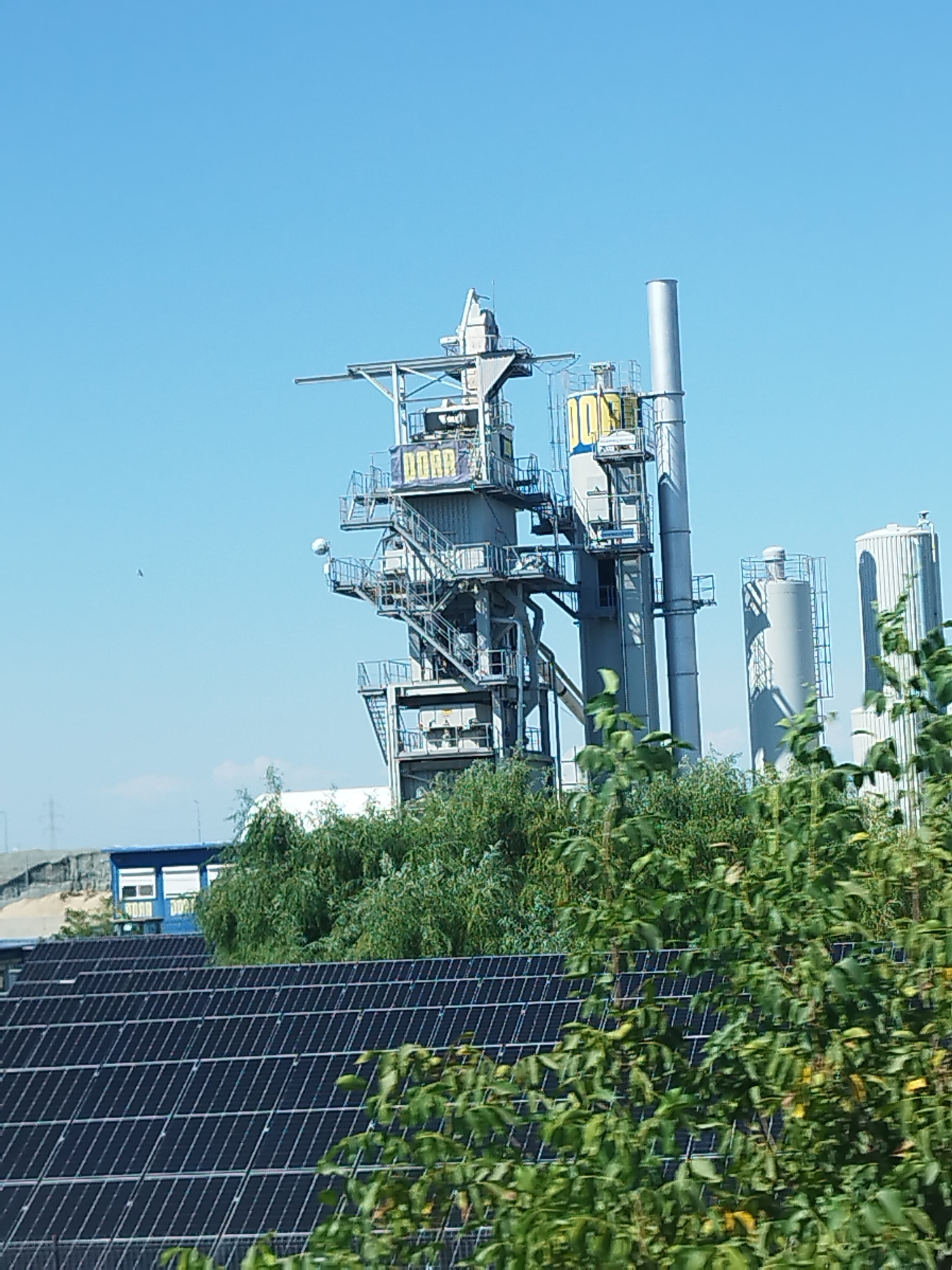 In Buftea, Romania, a modern industrial facility rises against a clear blue sky. The central structure is a complex of tall, gray metal towers and platforms, adorned with what appear to be large, yellow letters spelling out "DOAR" on one of the towers. Numerous staircases and walkways crisscross the structure, suggesting multiple levels and access points. Adjacent to this main tower are two large, cylindrical silos, also gray and metallic, one taller than the other. In the foreground, a vast expanse of dark solar panels covers a rooftop, suggesting a commitment to renewable energy. Lush green trees and foliage partially obscure the view of the industrial complex, creating a contrast between the natural and the manufactured. The scene is one of functional industry harmonizing with its environment, under a bright, sunny sky.