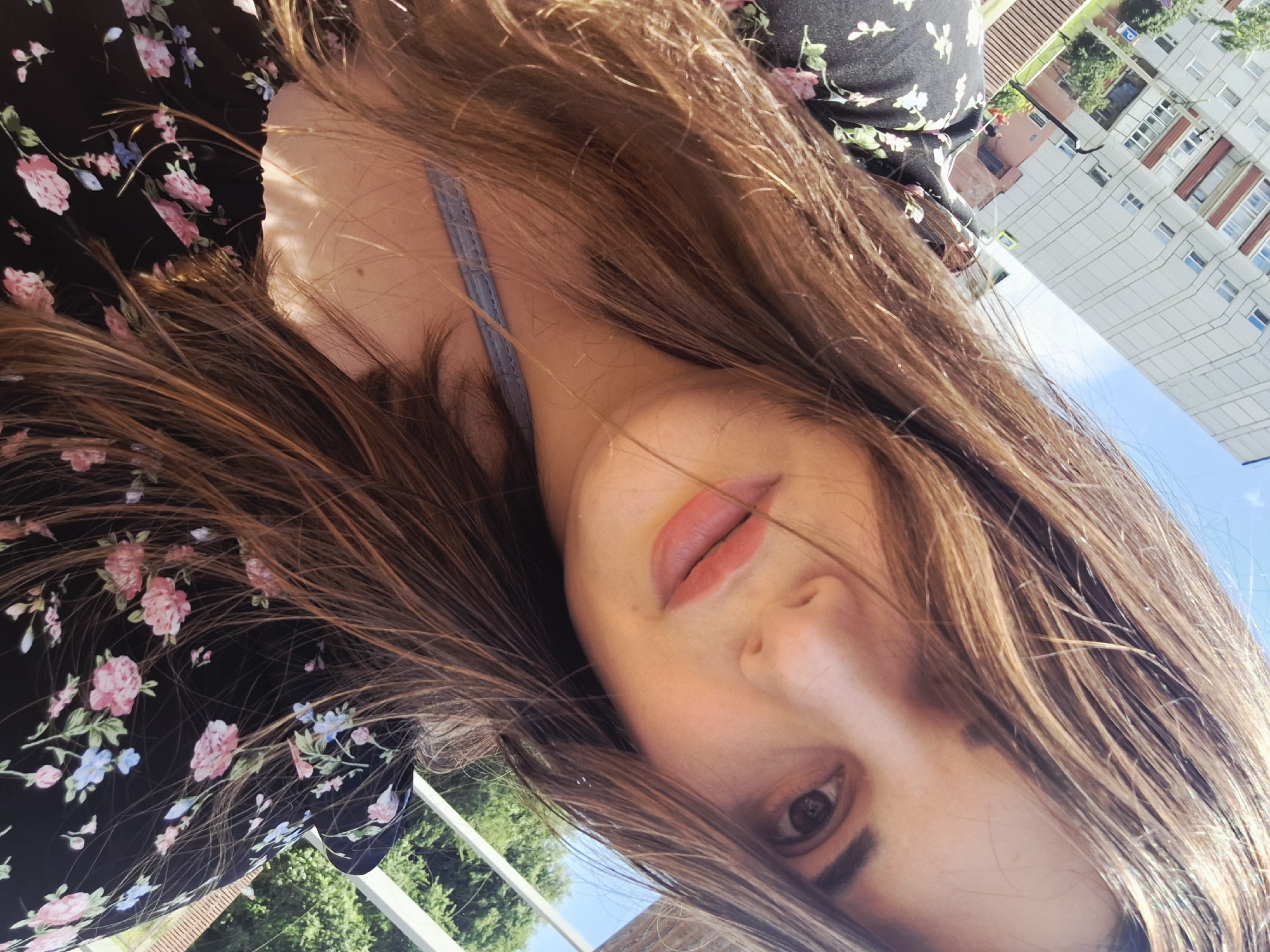 A young woman is taking a selfie outdoors. She has long brown hair, which partially obscures her face. Her head is tilted, and she is looking towards the camera. She is wearing a black dress with a floral pattern of pink and blue flowers. A light blue strap is visible across her chest. The background shows a residential building with a light-colored facade and balconies, a blue sky, and green trees. The scene appears to be set in a city environment, possibly in Chaikovsky, Russia, given the architectural style of the building.