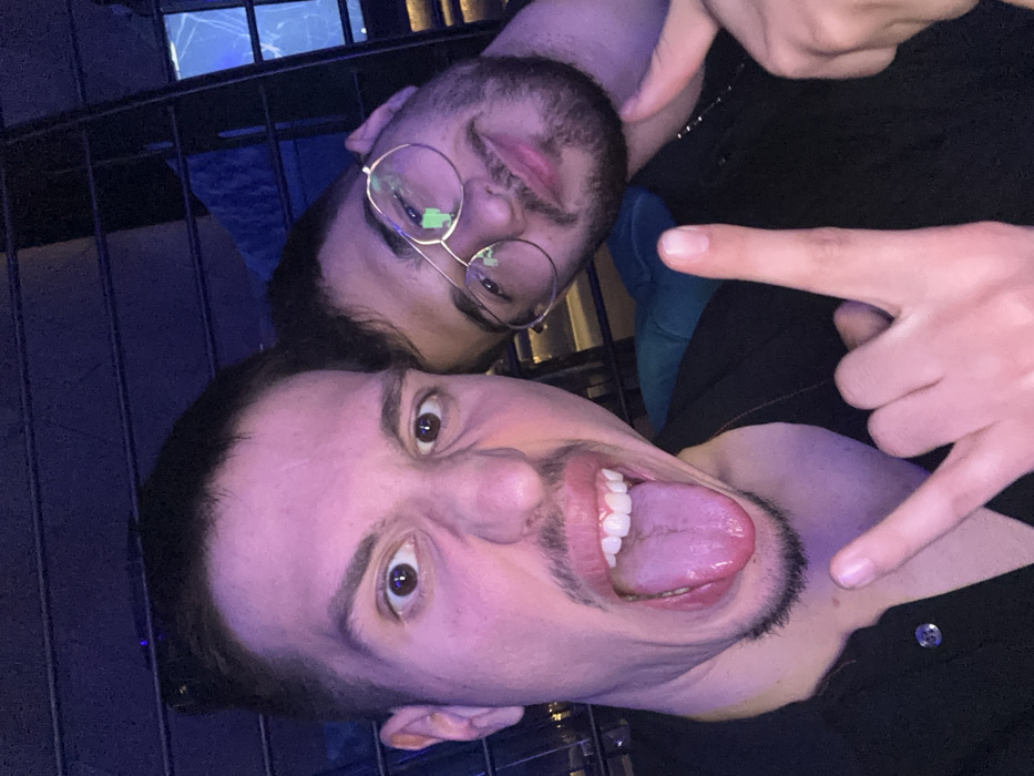 The image depicts two adult males posing for a selfie-style photograph in a dimly lit indoor environment, identified as a social venue within the city of Varna, Bulgaria. The background illumination features prominent blue and purple hues, characteristic of a bar or club setting.
The male in the foreground, positioned on the left side of the frame, has short dark hair, a short mustache, and a chin beard. He is actively making a humorous facial expression, with his tongue extended from his open mouth and his nose visibly scrunched, while his eyes are wide open and directed towards the camera. He wears a dark, collared shirt, open at the neckline. His left hand is raised in the lower-center, forming a "rock on" gesture with the index and pinky fingers extended.
The second male is situated behind and to the right of the first, his head slightly tilted. He has dark hair, a beard, and wears round, gold-rimmed spectacles that show reflections. He exhibits a subtle smile. His attire consists of a dark garment and a thin chain necklace. His right hand is visible in the lower-right corner, making a gesture with the thumb pointed upwards.
The background is largely out of focus, showing blurred dark structural elements, possibly railings or a partition, and abstract light patterns.