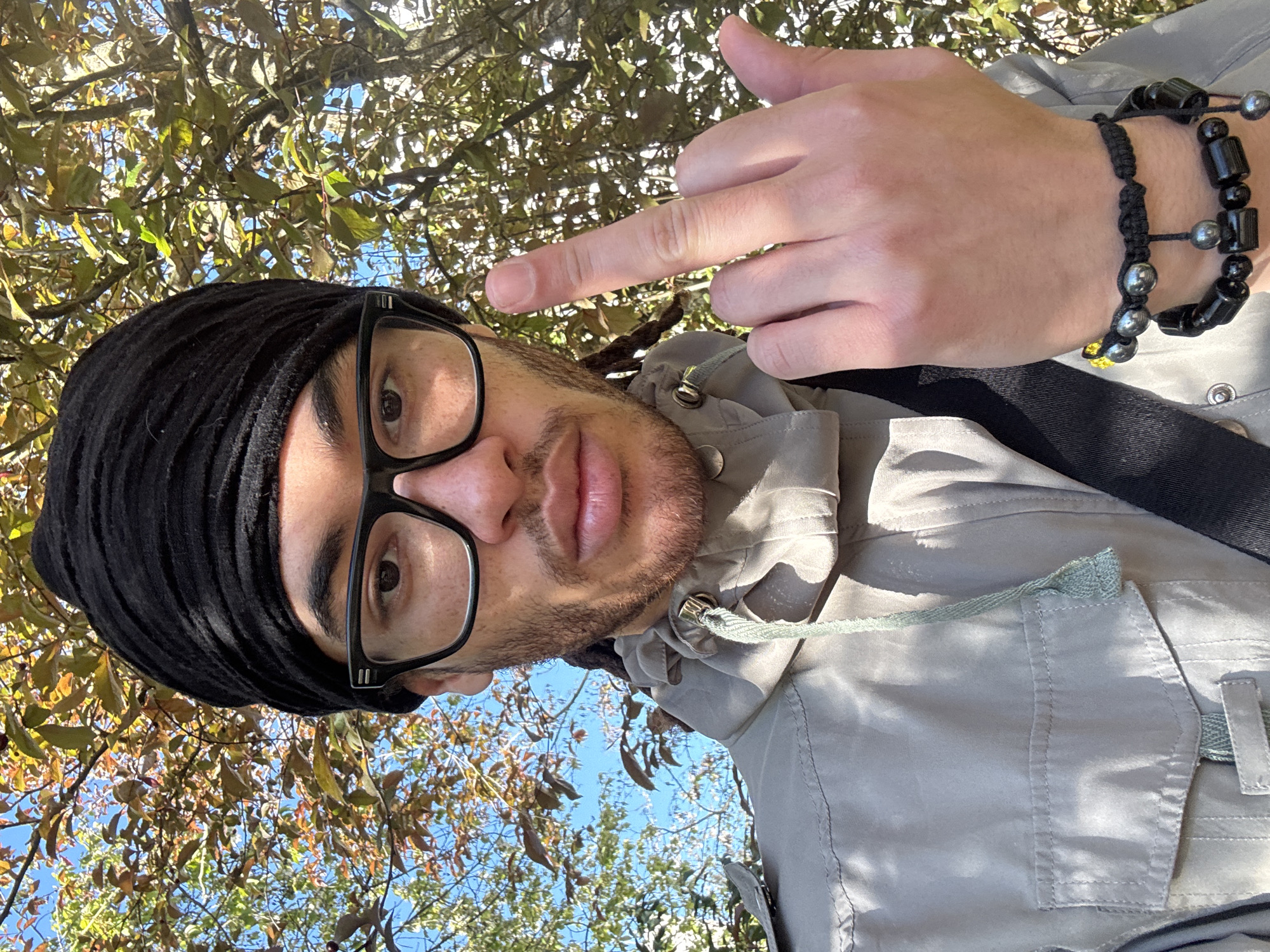 A selfie taken from a low angle shows a man wearing a black head covering and black-rimmed glasses. He is giving the middle finger. He is wearing a light grey jacket with a drawstring around the collar. He is wearing two black and grey bracelets on his right wrist. The background shows tree branches with leaves and blue sky. The location is most likely not Les Ulis, France as this cannot be determined from the image.