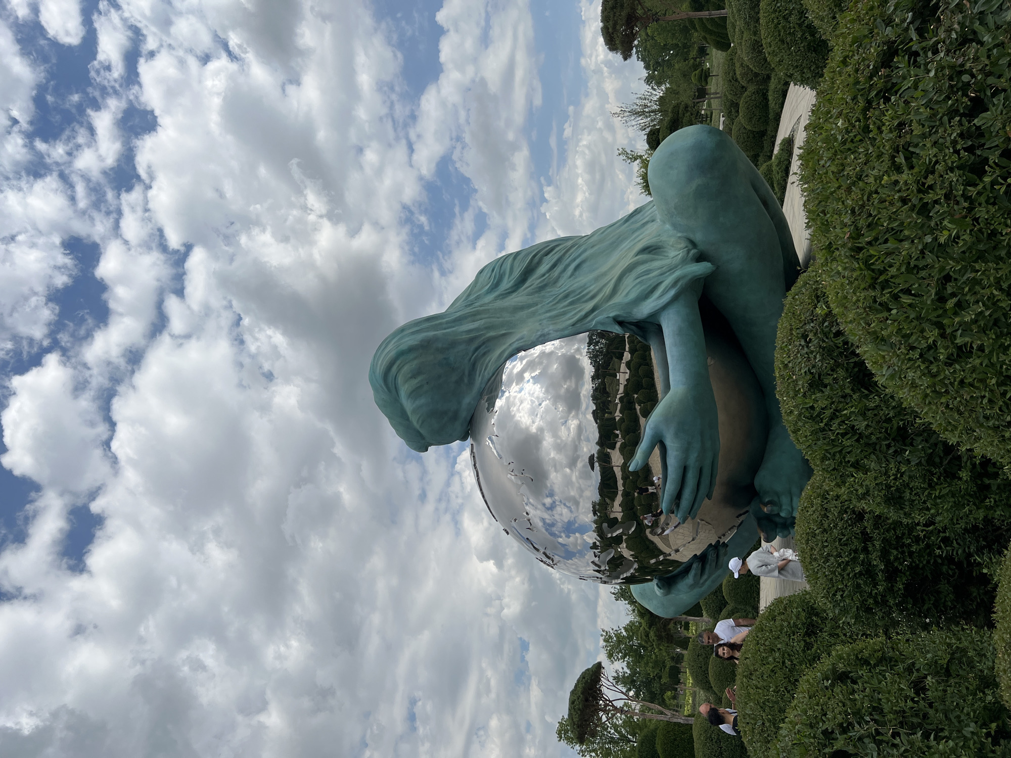 The image shows a large, teal-colored statue of a seated figure cradling a reflective sphere. The statue's hair flows around the sphere, and its hands are gently placed on it. The sphere reflects the sky, which is filled with white, fluffy clouds against a blue backdrop.
The statue is situated in a manicured garden with lush green bushes and trees. In the background, there are more sculpted bushes and what appears to be a pathway. Several people are visible in the background, some appearing to be tourists admiring the statue and the garden. One person in the middle ground is wearing a white cap and a light-colored outfit, possibly holding a baby. Other people are seen in smaller groups further in the background.
The lighting suggests it is daytime, likely late morning or afternoon, given the brightness and the shadows. The weather appears pleasant, with a partly cloudy sky. The overall scene evokes a sense of art appreciation and leisure in an outdoor environment. The location is identified as Krasnodar, Russia. No visible text is present in the image.