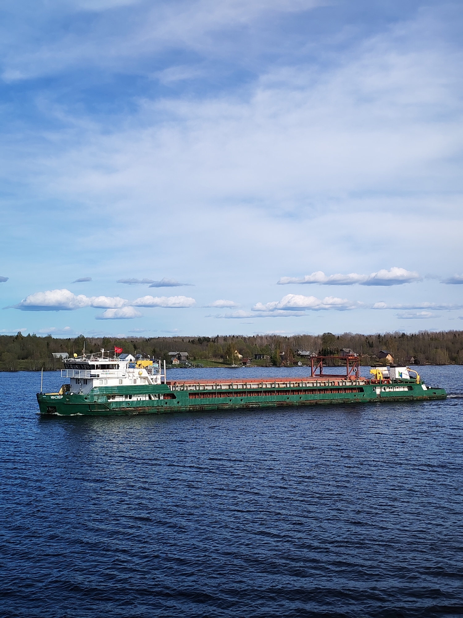 A long, green cargo barge labeled "ИНЗЕР" is moving across a body of blue water. The barge has a white cabin area on the left side, with various structures and what appears to be a red flag. The cargo area is filled with a reddish-orange material and includes a red crane structure. The background features a tree-lined shore with scattered buildings, suggesting a rural or semi-rural area. The sky is blue with scattered white clouds. The water has ripples suggesting movement. The location is likely near the city of Turtsevo, Russia, given the writing on the barge.