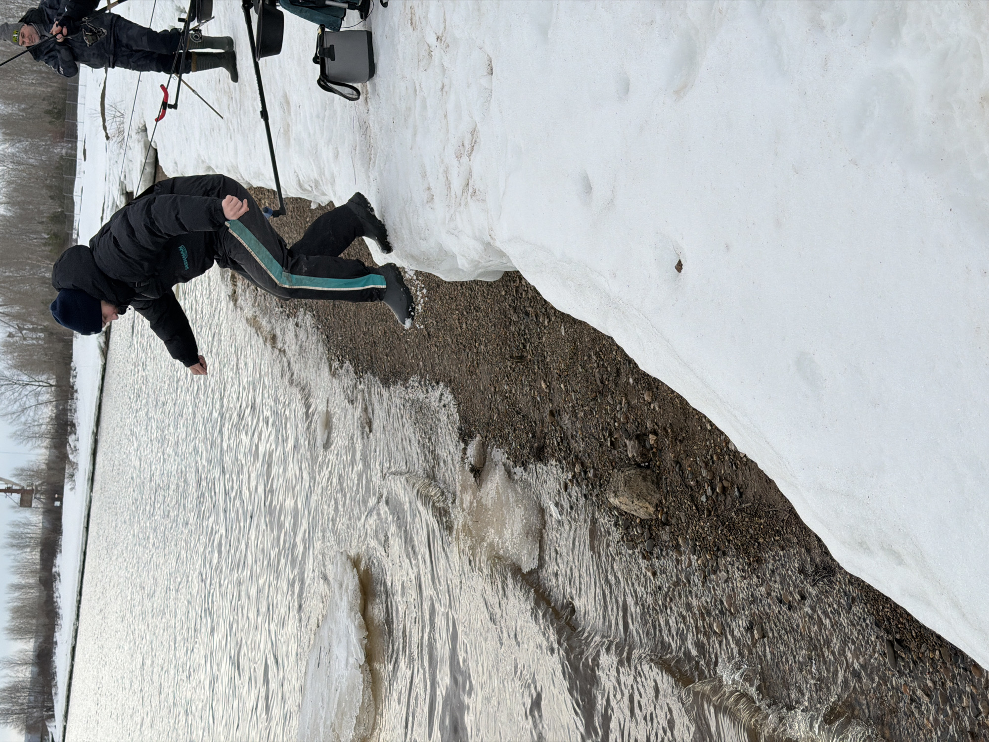 A view facing the edge of a body of water and land in Noviy, Russia. An individual wearing a black jacket, dark pants with light green side stripes, and a dark beanie is leaning over the water's edge. To the left of this person is the waterway with snow and ice chunks. A strip of land separates the water from a snow bank. Another person stands on the snow bank, dressed in dark clothing and holding fishing poles and gear. In the distance, the opposite shore can be seen lined with trees.