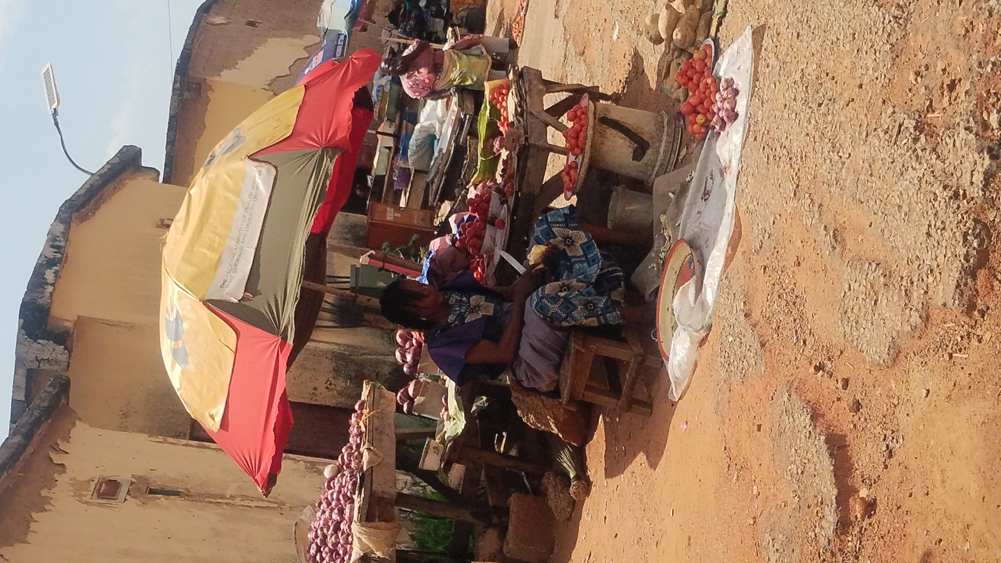 A slightly rotated outdoor shot shows an open-air market stall somewhere in the city of New Stadium, Nigeria, as inferred from the partially visible text "THE FEDERAL MINISTRY OF HEALTH WAR THAT SMOKERS ARE AS ARE LIABLE TO DUE YE" printed on an umbrella.
Two women, wearing what appears to be printed cotton clothes, are seated at the stall. The stall has several sections of produce on display, including tomatoes, onions, and what could be potatoes or yams. Some of the produce is arranged in piles. The foreground and ground are a light reddish-brown color.
The stall is in front of a building. A street lamp is attached to the building. The stall is shaded by a multi-colored umbrella, likely intended to provide protection from the sun.