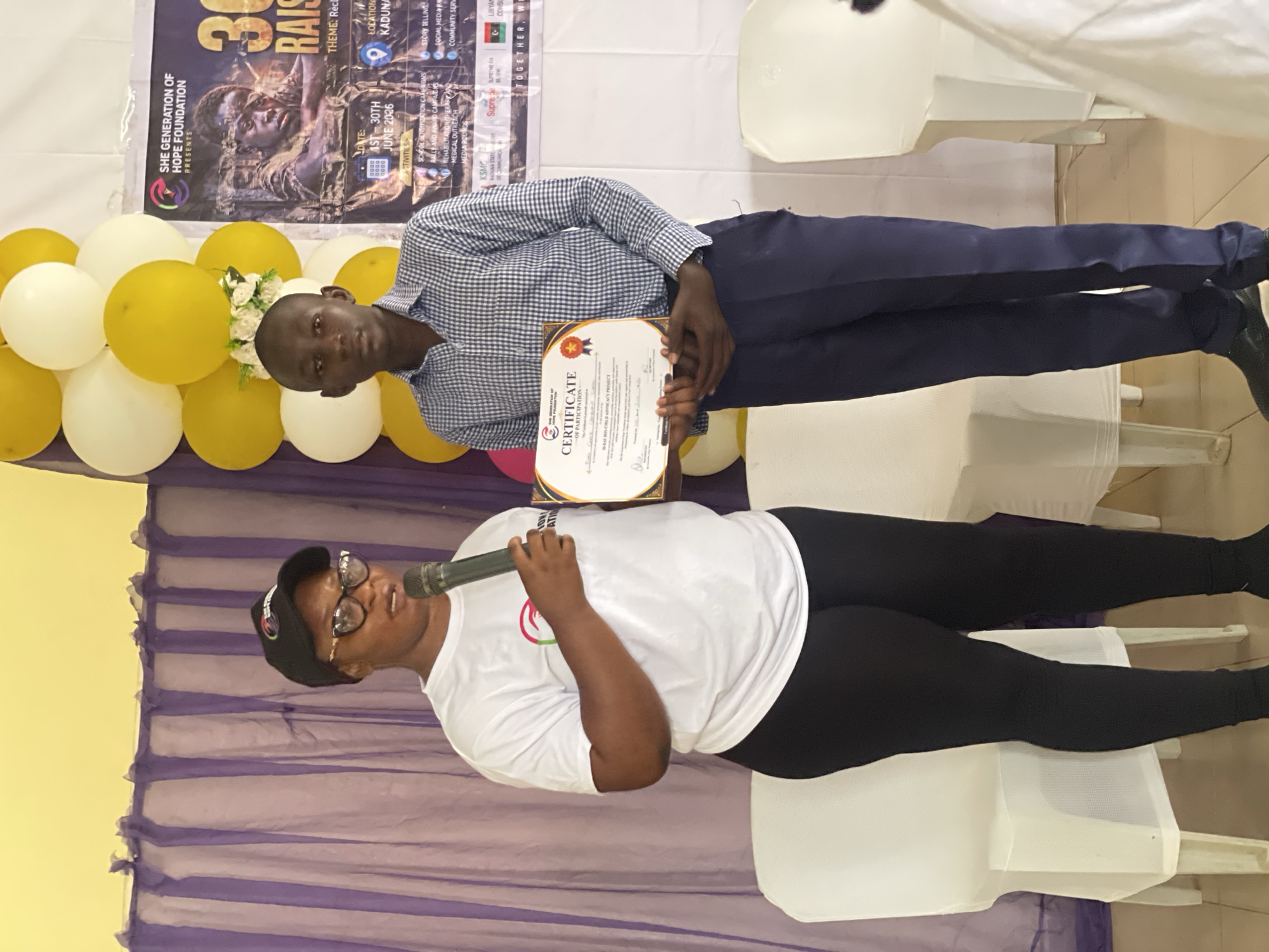 Two people are posing for a photo in front of a backdrop. The person on the left is a woman wearing glasses, a black baseball cap, a white t-shirt, and black leggings. She is holding a microphone and smiling. The person on the right is a man wearing a checkered blue and white shirt and dark blue pants. He is holding a certificate. In the background, there is a banner with text that includes "SHE GENERATION OF HOPE FOUNDATION PRESENTS 30 RAIS" and "THEME: Rec", "DATE: 1ST-30TH JUNE 2026", and "LOCATION: KADUNA". There are also balloons in yellow and white. The scene appears to be an indoor event. The overall impression is one of celebration or recognition, possibly an award ceremony or a community event held in Kaduna, Nigeria.