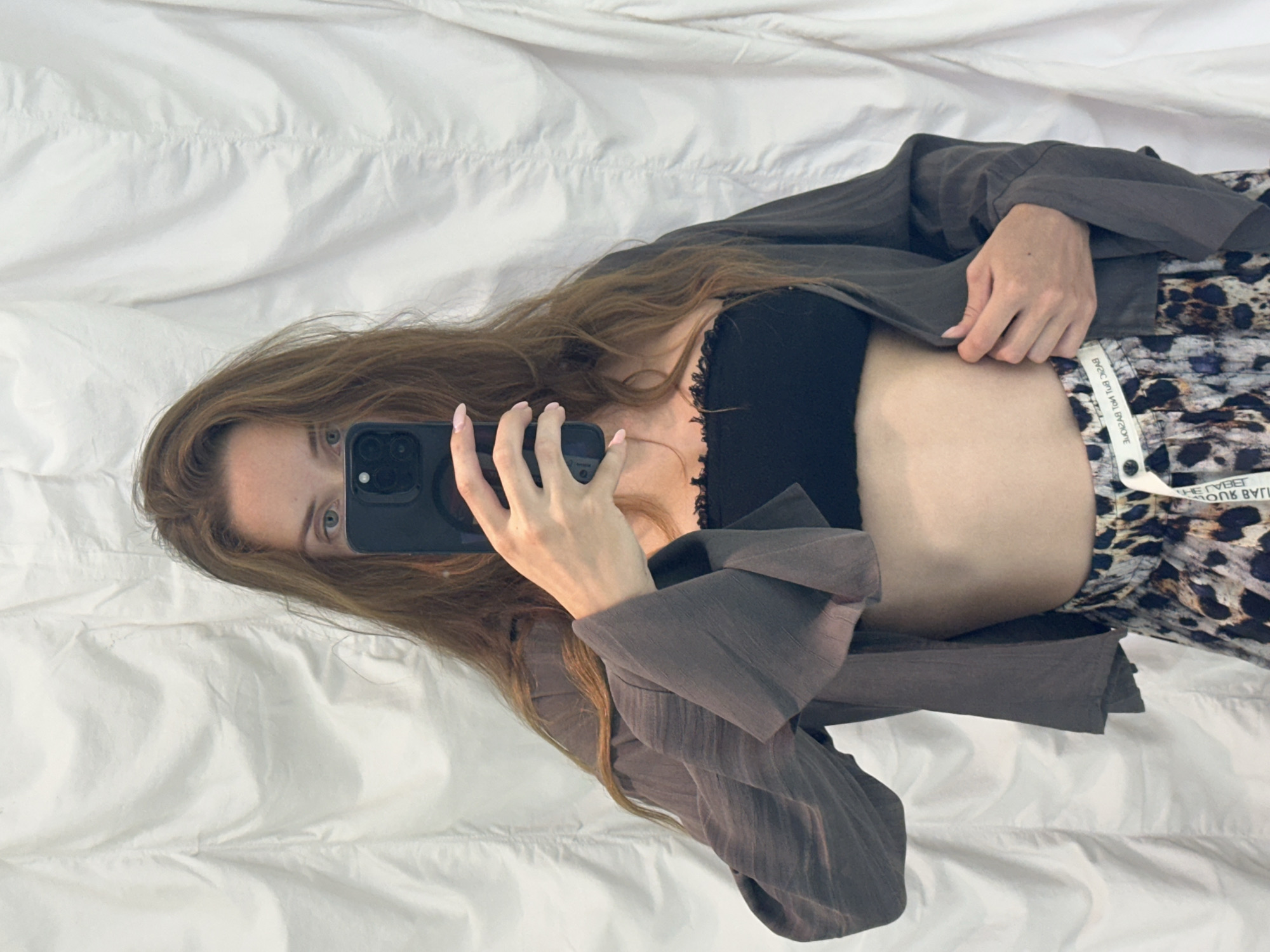 A female subject is depicted taking a mirror selfie in North Kuta, Indonesia. The subject has long, wavy, reddish-brown hair and light skin. Her face is partially obscured by a black smartphone, likely an iPhone, which she holds with her right hand; her left eye, which is blue-green, is visible. She wears a black strapless bandeau top with a ruffled lace trim along the upper edge, exposing her midriff. Over the bandeau, she wears an open, dark grey, lightweight, long-sleeved shirt with wide cuffs. Her left hand is positioned near her waist, holding the edge of the grey shirt open. She wears loose-fitting trousers or shorts with an abstract, multi-tonal animal or camouflage print in shades of brown, black, and grey. A white tag or drawstring, partially inscribed with reversed text including "BASIC BUT NOT BASIC" and "THE LABEL," is visible on the waistband of her bottoms. The background consists of a plain white or off-white textured fabric surface with vertical folds.