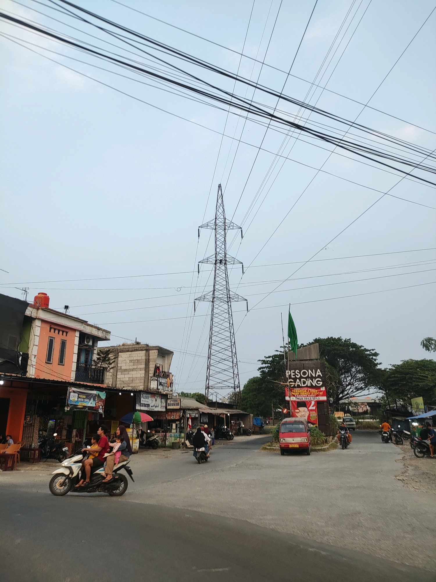The image depicts a street scene in Wanajaya, Indonesia, characterized by a prominent electricity pylon and overhead power lines. On the left side of the street, there are several buildings, including a two-story structure painted orange and white, with a red water tank on its roof. Adjacent to it are other buildings constructed with concrete blocks and some covered with tarpaulin. A few people are visible, with one motorcycle carrying a family of four riding on the road. Several other motorcycles are parked or being ridden.
In the center of the image, the tall electricity pylon dominates the skyline. To the right of the pylon, a green flag is mounted on a wooden sign that reads "PESONA GADING". A small red vehicle, possibly a minivan, is parked near the sign. Further down the road, more buildings and trees are visible, along with several individuals and motorcycles. The sky is overcast, suggesting a cloudy day. The road surface appears to be asphalt.