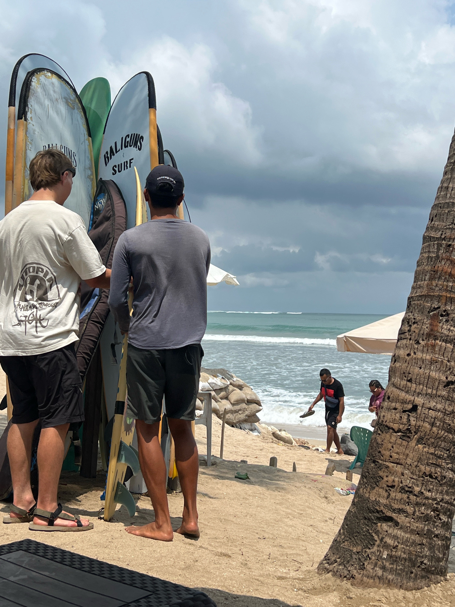 The image depicts a sandy beach in Kuta, Indonesia, with the ocean and cloudy sky in the background. In the foreground, a collection of surfboards stands upright, leaning against each other. Two young men, viewed from behind, are standing next to the surfboards. One is wearing a light-colored t-shirt with a graphic print and black shorts, and the other is in a dark long-sleeved shirt and dark shorts. Further down the beach, a man in a black t-shirt and shorts is walking towards the water, holding a flip-flop. A woman in a purple top is standing near him. Beach umbrellas are visible further in the background, and a portion of a palm tree trunk is on the right side of the frame.