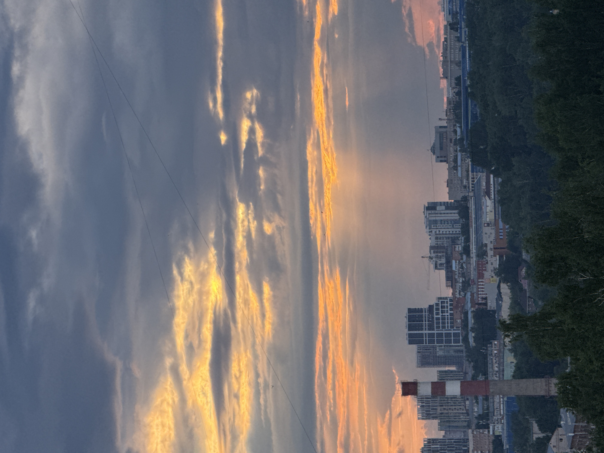The image captures a vibrant cityscape under a dramatic sunset or sunrise sky, presumably in Izhevsk, Russia. The sky dominates the upper portion of the frame, painted with striking contrasts. On the right, elongated streaks of clouds glow intensely with warm orange and yellow hues, catching the direct light of the sun, which appears to be setting or rising off-frame to the right. These bright clouds transition into a darker, cooler palette of grey and blue clouds on the left, creating a strong atmospheric effect. Thin, dark utility wires or power lines are faintly visible stretching across the sky.
Below the horizon, a sprawling urban landscape unfolds. A mixture of residential and commercial buildings forms a dense skyline, with several multi-story modern structures and older buildings discernible. In the foreground on the right side of the cityscape, a prominent industrial chimney stands tall, distinguished by its red and white striped pattern. A lush, dark green line of trees or forest occupies the lower right and central background, suggesting a natural boundary or parkland bordering the city. No specific human activity is overtly visible, but the scene portrays the serene beauty of an urban environment at dusk or dawn. No discernible text is visible within the image.